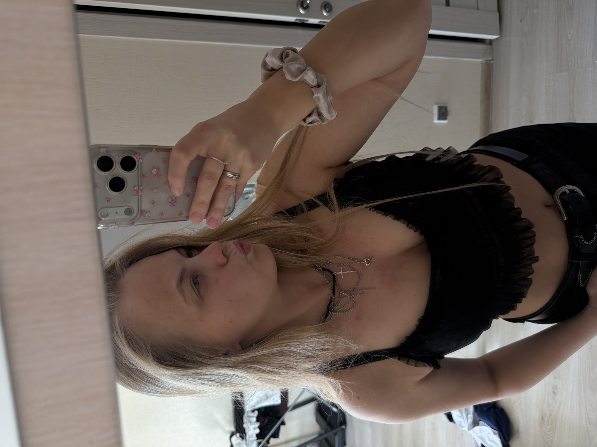 A mirror selfie, originally oriented on its side, captures a young woman indoors. She has long, straight blonde hair and is looking towards the camera with a slight pout. Her complexion is fair, with some minor blemishes visible on her face.
She is wearing a black top with ruffled detailing around the neckline or bust, and a black belt with a silver buckle over dark-colored bottoms. Several silver necklaces adorn her neck, including one with a prominent cross pendant and another with a small heart charm. On her left hand, she wears two rings: a simple silver band on her ring finger and a more ornate one on her middle finger. A light beige or silver scrunchie is worn on her right wrist.
The woman is holding a smartphone in a clear case decorated with small pink floral patterns. The phone's triple-lens camera system is clearly visible in the reflection.
The background, reflected in the mirror, indicates an indoor domestic setting. To the left, light-colored wooden furniture, possibly a wardrobe or door, is visible. The wall behind her appears light and plain. The floor is also made of light-colored wood. In the lower right portion of the image (when oriented correctly), some folded or discarded clothing items, including white and dark blue fabrics, are visible on the floor, adding to the casual home atmosphere. There is no visible text in the image.