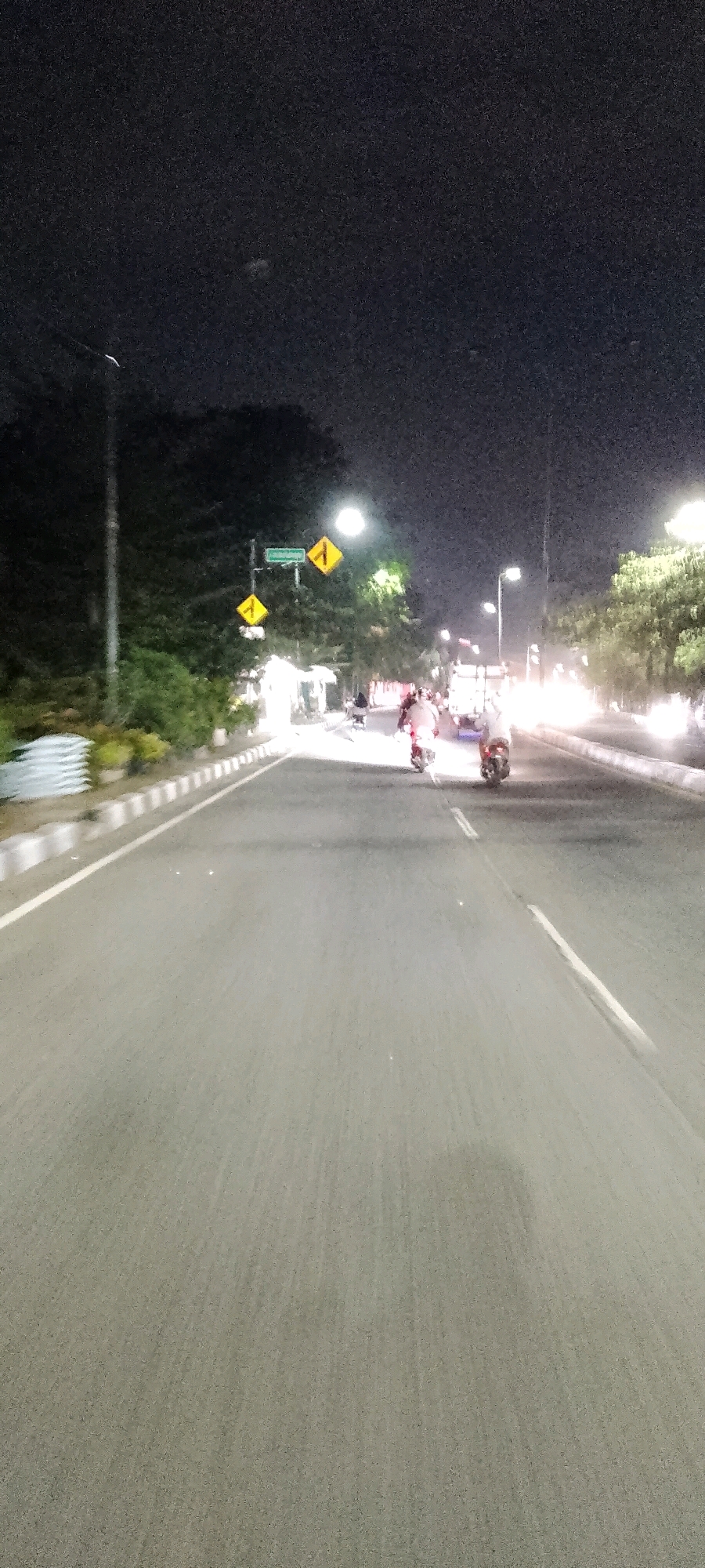 This is a nighttime, vertical, blurry photograph taken from the perspective of someone on a moving vehicle on a paved road. The road is asphalt with white lane markings and a white curb on the left side. Trees and foliage line the left side of the road, and there are streetlights visible. On the left side of the road, there are at least two yellow diamond-shaped traffic signs with black arrows. One sign indicates a turn to the left, and another indicates a change in direction with a left turn. There is also a green street sign.
In the middle distance, on the road, two individuals are riding motorcycles. The motorcycle on the right is closer to the center of the road, and the motorcycle on the left is further to the right. Both riders are facing away from the camera, and the headlights and taillights of the motorcycles are visible. In the background, there are more trees and lights, suggesting an urban or suburban environment. The overall impression is of a typical night scene on a road in Gunung Putri, Indonesia, possibly a main thoroughfare, with traffic and street lighting.