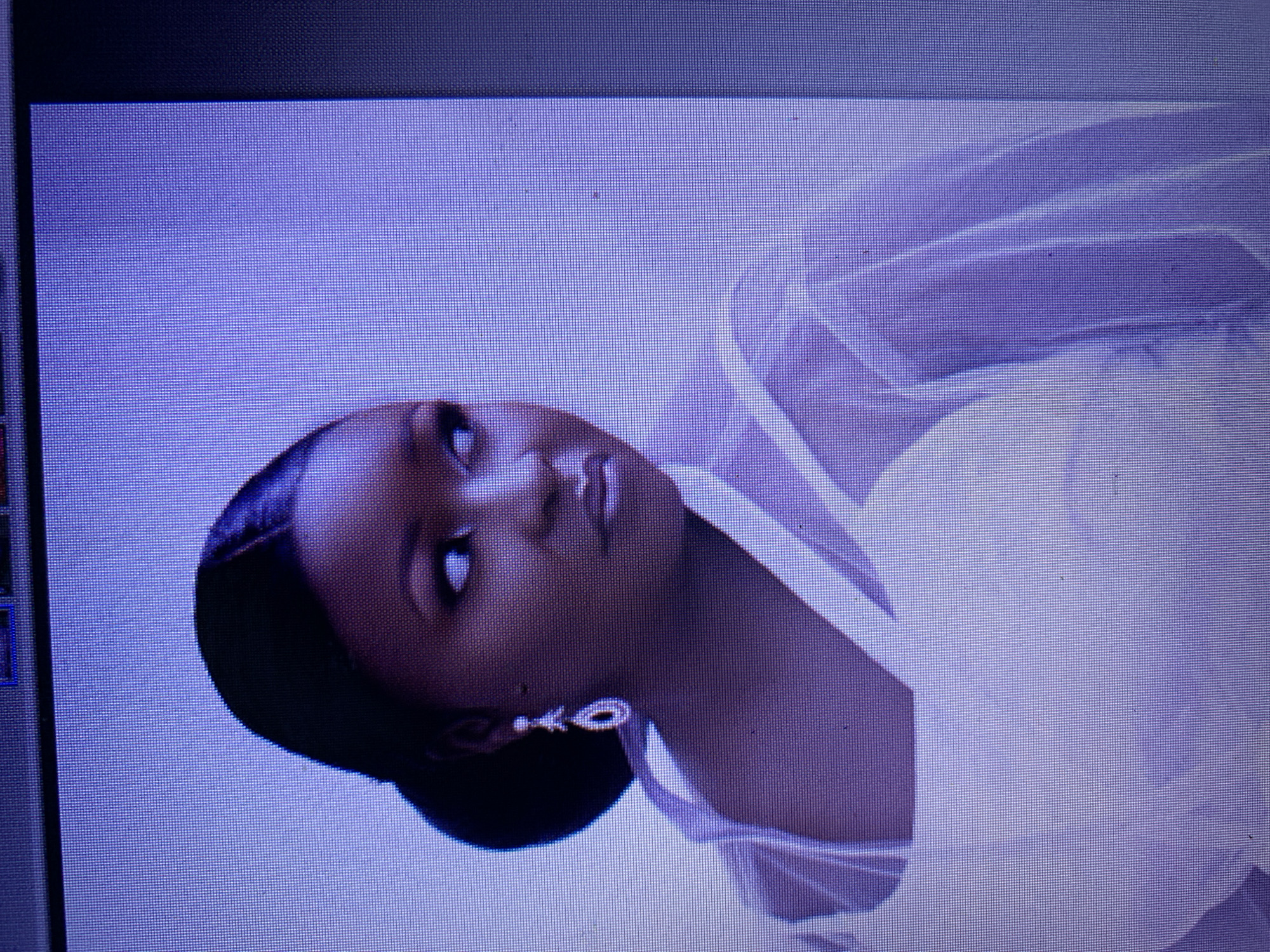 The image shows a close-up of a woman with dark skin and dark hair styled in a bun. She is wearing a white, sheer garment and a teardrop-shaped earring. Her head is turned to the right, and she is looking off into the distance with her eyes wide. The background is a solid light purple color. There is no discernible indication of the city of Jos, Nigeria, or any other specific location. The image appears to be a studio portrait.
