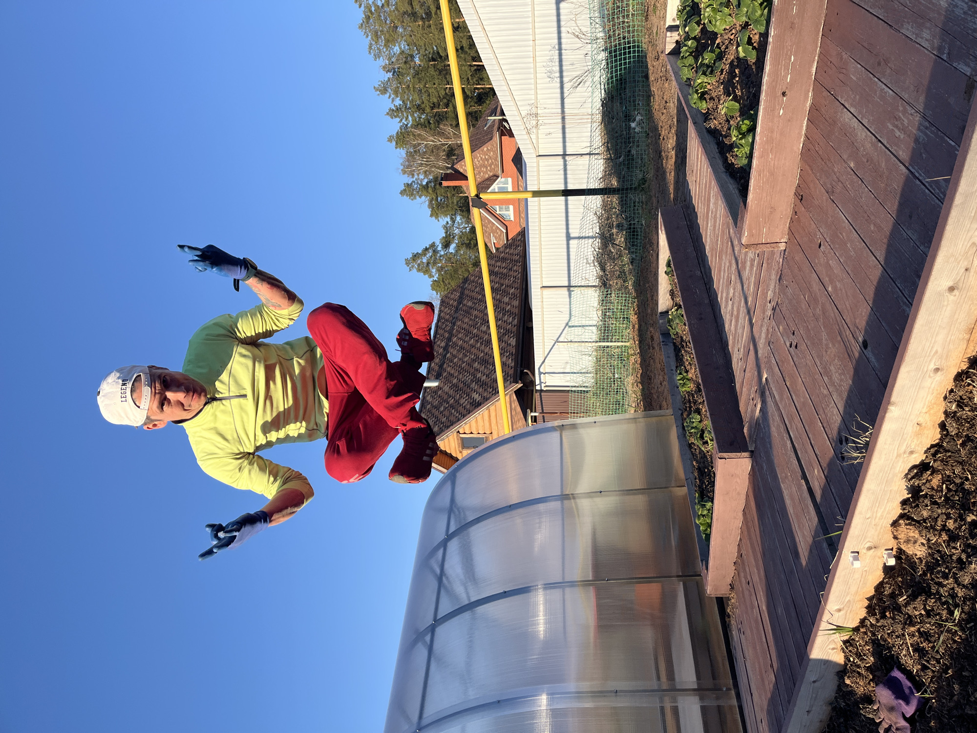 A man is photographed in what seems to be a staged, playful pose in a yard. He is wearing a light green long-sleeved shirt, red pants and shoes, a white cap with the word "LEGEND" on it, and blue gloves. He is positioned above a clear arched structure that could be a greenhouse or a tunnel, with a blue sky visible behind him. The background includes a fence, trees, and a building. The area also features a wooden walkway or structure next to a patch of dark soil. The location is Chaikovsky, Russia.