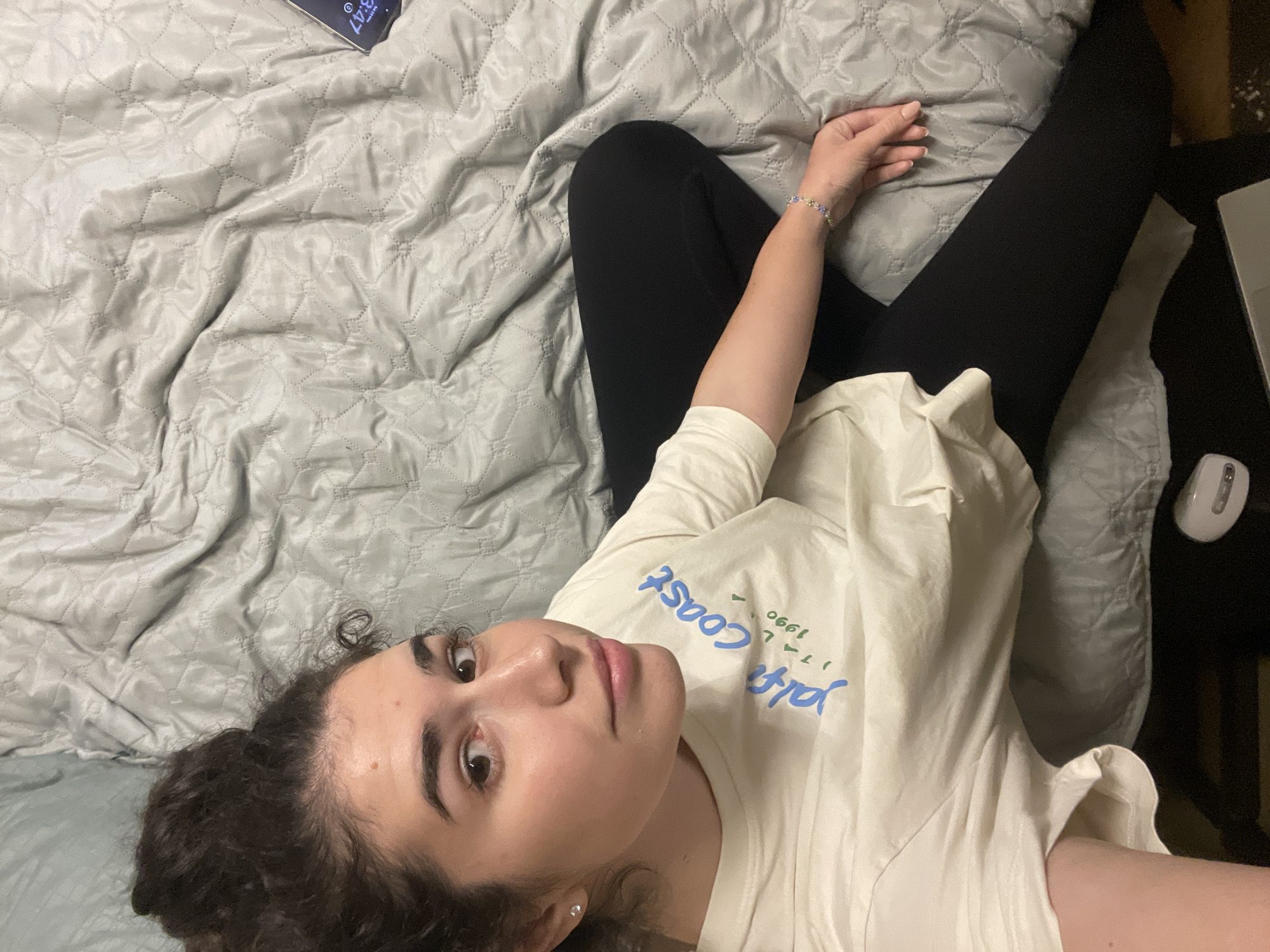 A young woman with curly brown hair is lying on a bed covered with a light gray patterned quilt. She is wearing black leggings and a light-colored t-shirt with blue and green lettering. Her right arm is extended on the bed, and she is wearing a bracelet on her wrist. A laptop and a wireless mouse are on a dark surface, possibly a nightstand, to the right of her. The image was likely taken indoors. The location is not determinable from the provided image.