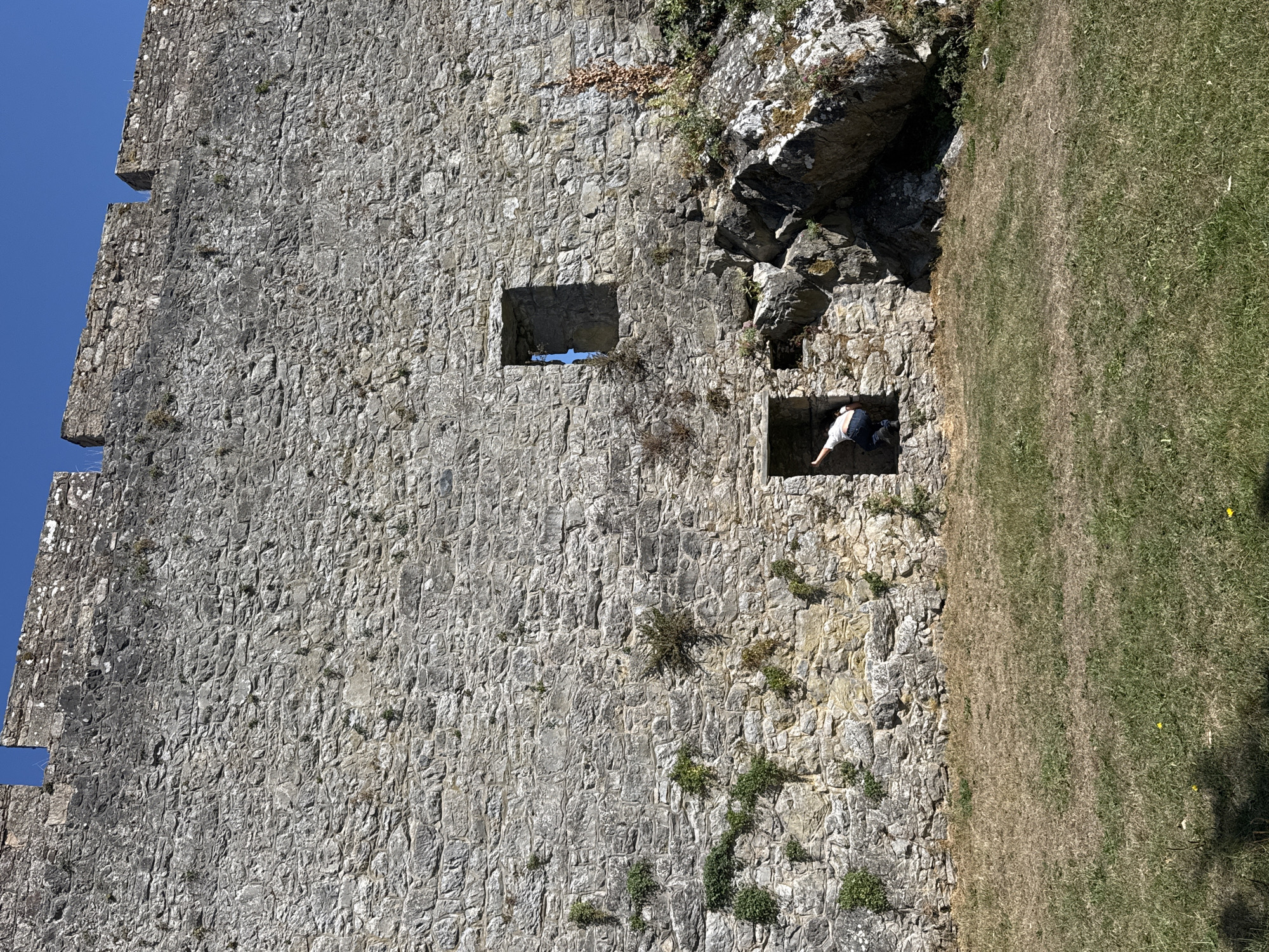 A person in a white shirt and blue jeans is climbing out of a small, stone opening in a large, ancient stone castle wall. The castle wall is made of rough, grey stones and has some small green plants growing on it. There are two openings in the wall: one larger, rectangular opening higher up and a smaller, square-shaped opening where the person is. Below the wall, there is a grassy area with some dry patches and a few small yellow flowers. To the right of the person, large rocks are embedded in the ground against the castle wall. The sky is clear and blue. This scene is located at Cahir Castle, in Cahir, Ireland.