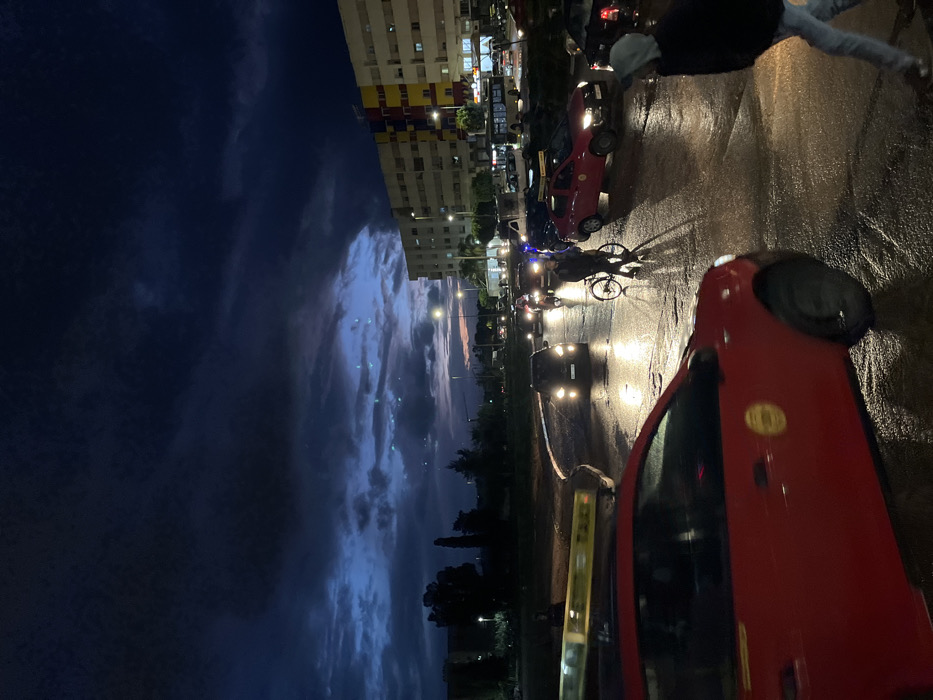 A street scene at night features traffic including cars and a cyclist. Headlights illuminate the wet road surface. A multi-story building is visible along with silhouetted trees under a cloudy sky. One red car is prominent in the foreground.