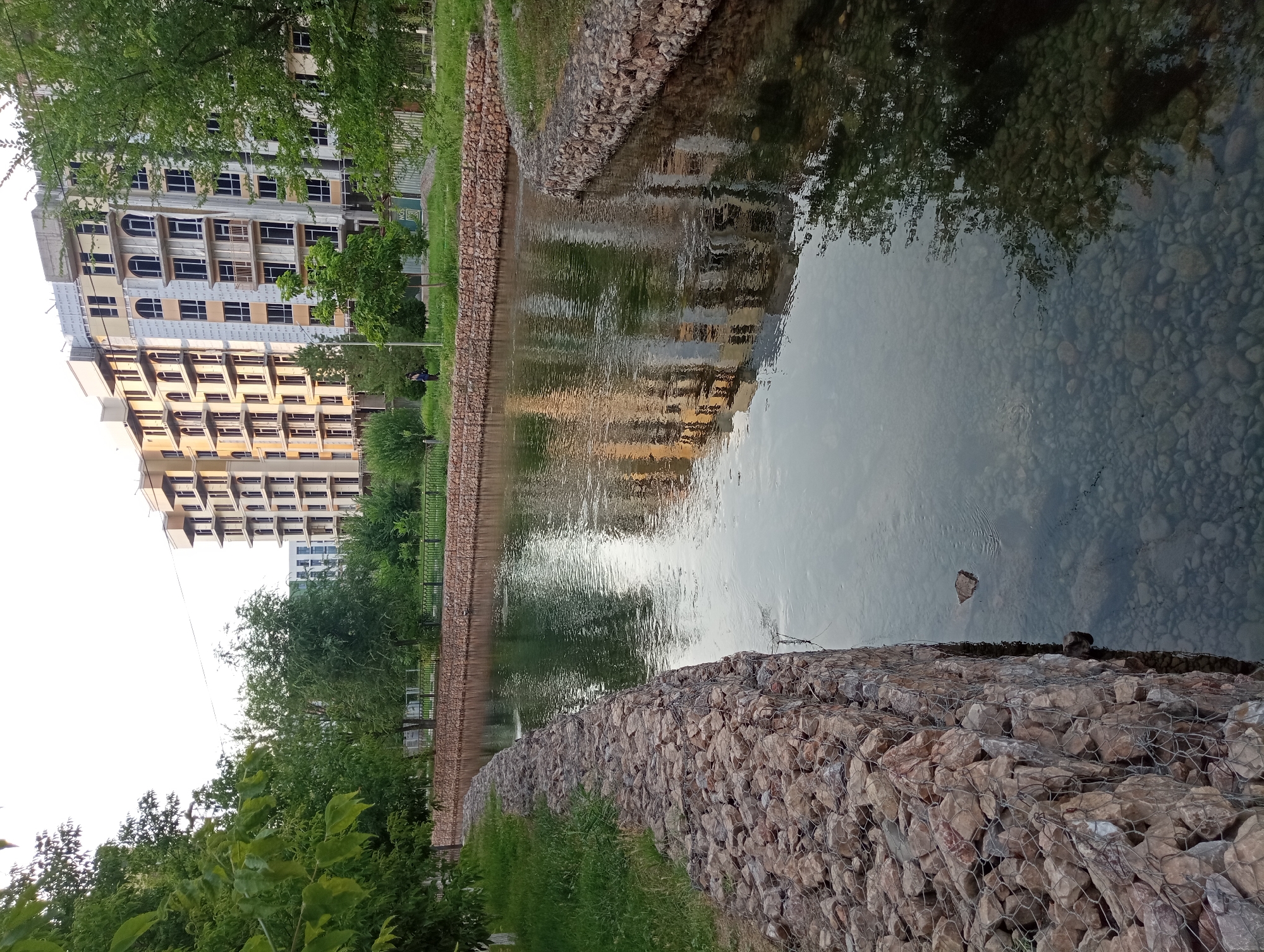 The image depicts a canal with clear water, lined by gabion walls constructed of large stones and wire mesh. The canal runs through a park-like setting with lush green grass and trees on either side. In the background, a multi-story building, under construction, dominates the skyline. The building's facade shows exposed concrete and scaffolding, indicating ongoing construction work. The water reflects the building and the surrounding trees. A single person, appearing as a silhouette from this distance, is visible on the grassy bank in the middle ground. The scene is illuminated by natural light, suggesting it was taken during the daytime. This area is located in the city of Shymkent, Kazakhstan.