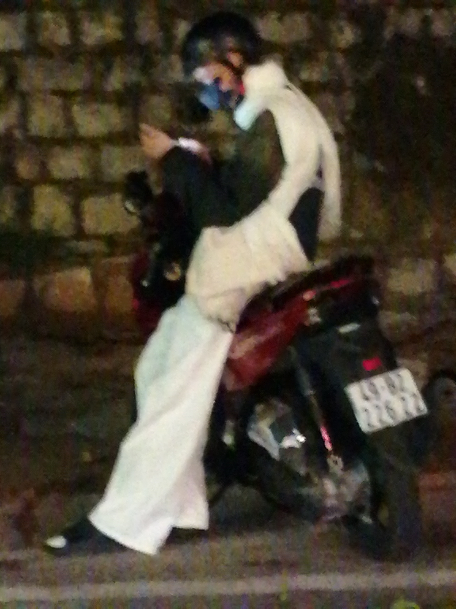 An out-of-focus, night time image shows a person sitting on a red and black motorcycle. The person is wearing a white garment, dark pants, and a dark helmet. They are holding a dark-colored mobile phone. A license plate with Vietnamese characters and numbers is visible on the back of the motorcycle. The background is a textured brick wall and the ground appears to be paved. The scene is likely in Da Lat, Vietnam, given the style of the license plate.