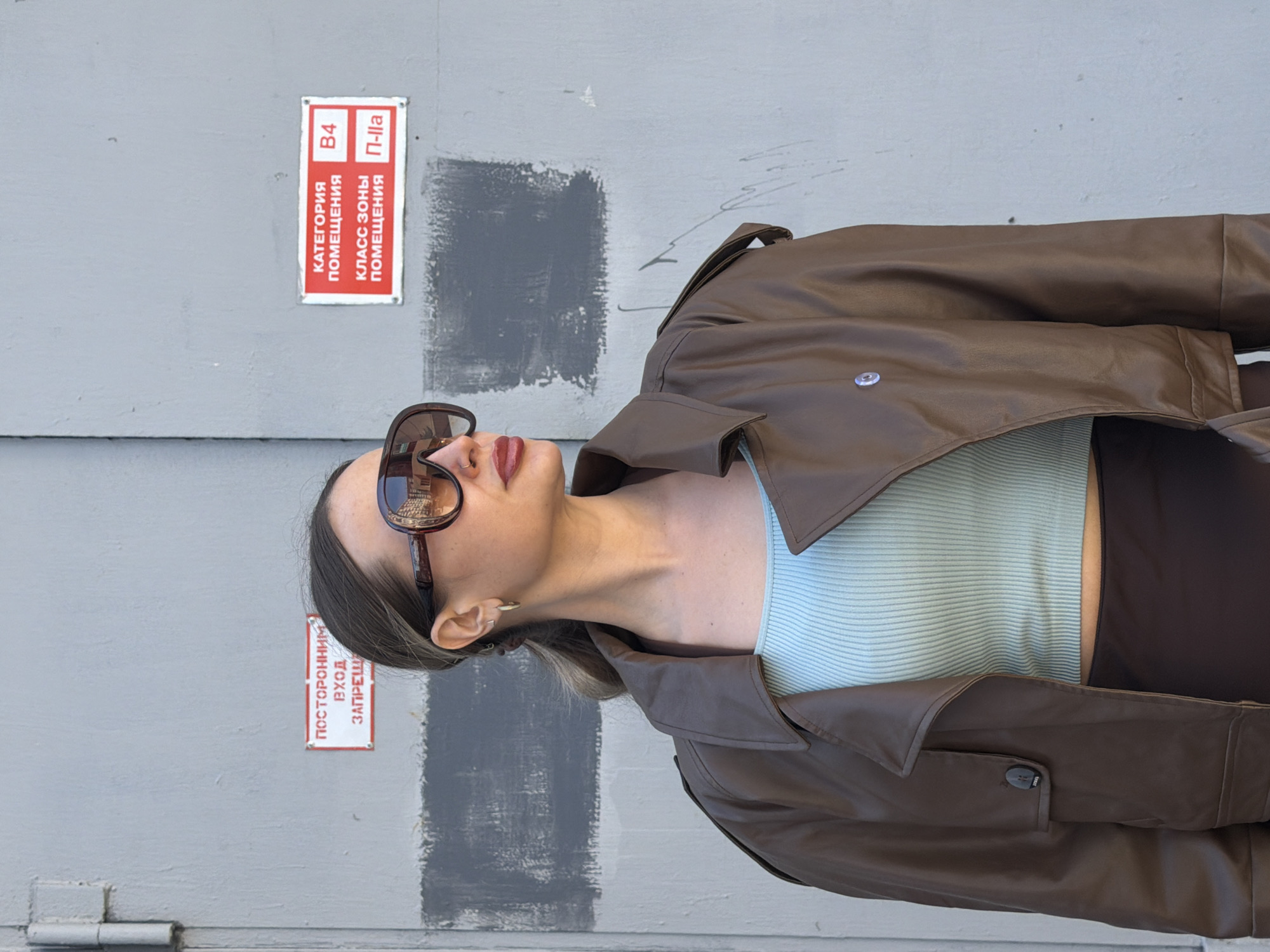 The image displays a woman posing for a photograph, viewed from the chest up, against a light grey wall. Her head is tilted slightly upwards and to her right. She is wearing large, brown, reflective sunglasses, a stylish brown faux-leather trench coat, and a light blue ribbed crop top underneath. Her dark hair is styled back, revealing a small hoop earring and a nose piercing. Her lips are adorned with reddish-brown lipstick.
The background consists of a plain, light grey wall, which appears to be made of metal or concrete. It features some darker grey painted patches and faint scuff marks. A metal hinge is visible in the bottom left corner, suggesting the wall may be part of a large door or panel.
Two red and white rectangular signs with Russian text are attached to the wall. The upper sign, prominently displayed, reads: "КАТЕГОРИЯ ПОМЕЩЕНИЯ" (CATEGORY OF PREMISES) "B4", and "КЛАСС ЗОНЫ ПОМЕЩЕНИЯ" (HAZARD CLASS OF PREMISES) "П-IIа". The lower sign, partially obscured by the woman's hair, states: "ПОСТОРОННИМ ВХОД ЗАПРЕЩЕН" (UNAUTHORIZED ENTRY FORBIDDEN).
The lighting is soft and even, characteristic of daytime under overcast skies or open shade, with no harsh shadows. The woman's posed stance and fashionable clothing suggest a deliberate photoshoot. The signs with Russian text confirm the location is within a Russian-speaking region, specifically Krasnoyarsk, Russia. Her attire, including the trench coat, suggests a mild to cool temperature.