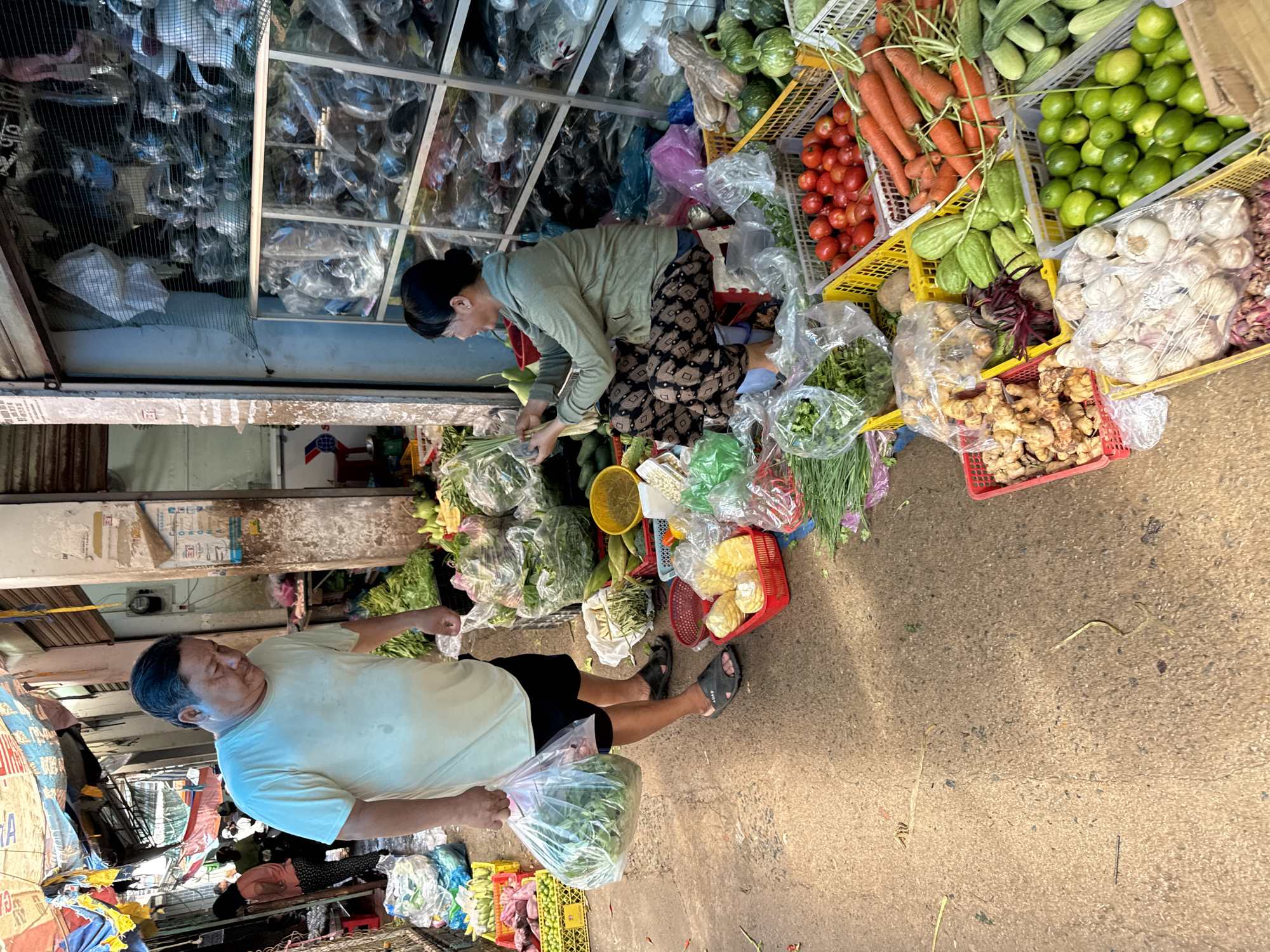 In Mũi Né, Vietnam, an outdoor market scene unfolds with a focus on fresh produce. A middle-aged man, wearing a light-colored t-shirt and black shorts, holds a clear plastic bag filled with green leafy vegetables. He is positioned on the left side of the image, facing away from the viewer and towards a woman. The woman, also middle-aged, is seated on a red stool in front of a stall stocked with various goods. She is wearing a green jacket and patterned pants, and is actively sorting through a bunch of green stalks. The market appears to be bustling, with stalls set up along a pathway. Crates and baskets overflow with colorful fruits and vegetables, including tomatoes, carrots, cucumbers, limes, garlic, ginger, and corn. Tarps and makeshift awnings provide shade, and the ground is a rough, paved surface. The overall impression is one of a vibrant, local marketplace dedicated to the sale of fresh produce.