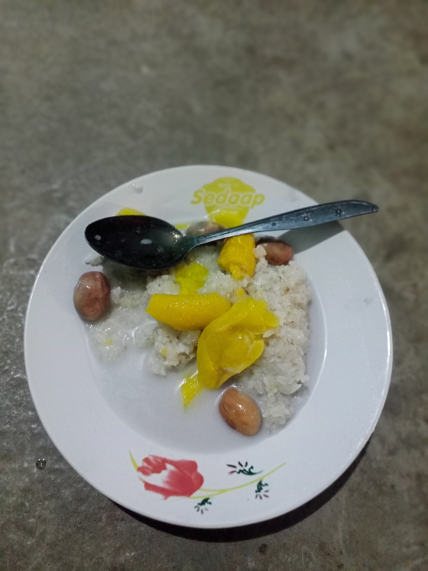 A plate of sticky rice with jackfruit and peanuts is shown, with a spoon resting on it. The plate is white with a floral design and the word "Sedaap" printed on it. The food appears to be in a dish of coconut milk. The plate is placed on a textured surface, possibly a table or countertop. The lighting suggests the image was taken indoors or at night.