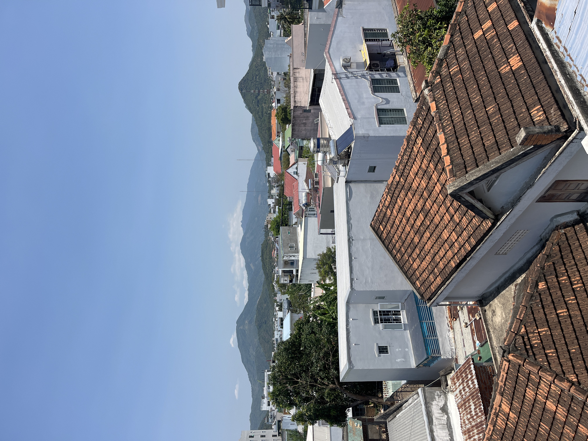 The image captures an aerial view of Bắc Nha Trang, Vietnam, characterized by residential buildings with terracotta tile roofs. The foreground is dominated by these roofs, casting shadows. Midground comprises dense clusters of low-rise structures, transitioning into the background where mountains ascend to meet a pale blue sky. No people are visible in the scene.