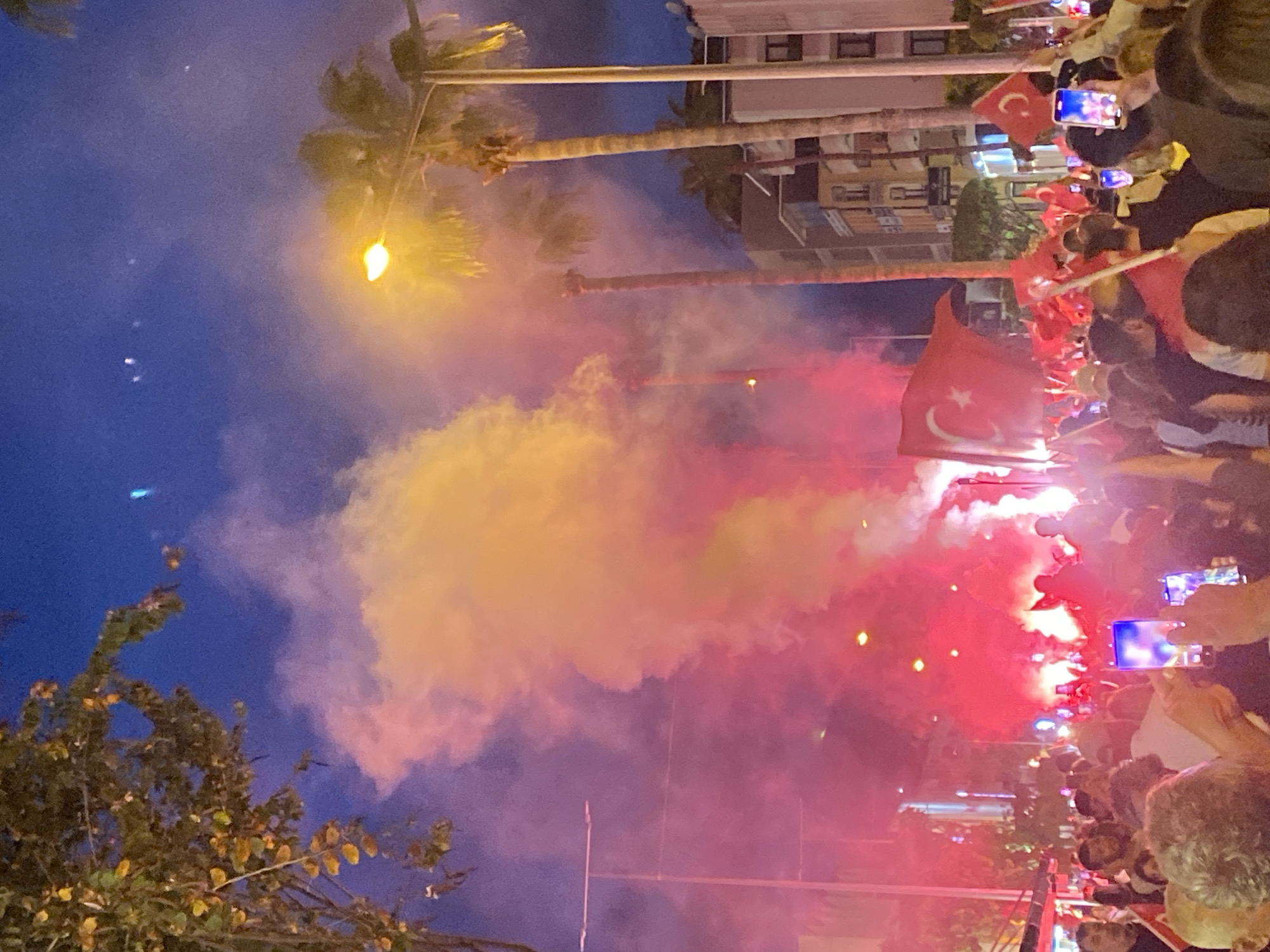 A low-angle view oriented vertically captures a nighttime celebration scene, likely in Alanya, Turkey. A crowd of people, many holding small Turkish flags, is visible, with some individuals filming the event on their phones. Red and yellow smoke fills the air, likely from flares or pyrotechnics. In the background, palm trees and buildings are visible, indicating an urban environment. A street lamp illuminates the scene. The sky is a deep blue, suggesting it is late evening. The focus is on the festive atmosphere, with the flags, smoke, and lights creating a vibrant spectacle.