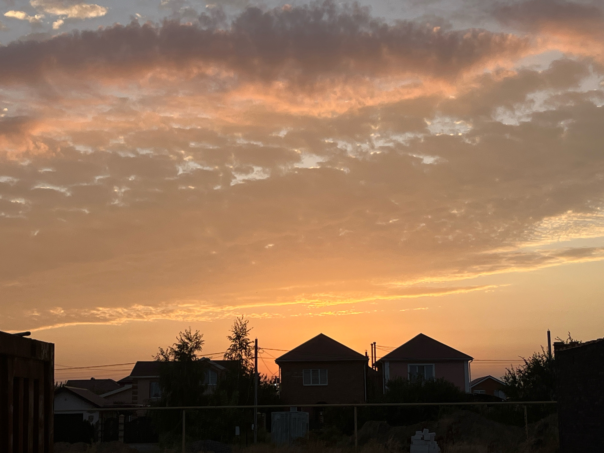 A wide-angle, eye-level shot captures a sunset over a residential area in Taganrog City District, Russia. The sky is dominated by a gradient of warm oranges and pinks, with scattered clouds tinged with the same hues. In the foreground, the silhouetted roofs of several houses are visible, partially obscured by dark trees and foliage. A chain-link fence runs horizontally across the lower portion of the image. To the far left, a dark, industrial-looking structure is partially visible. The overall impression is one of a quiet evening in a suburban or rural setting.