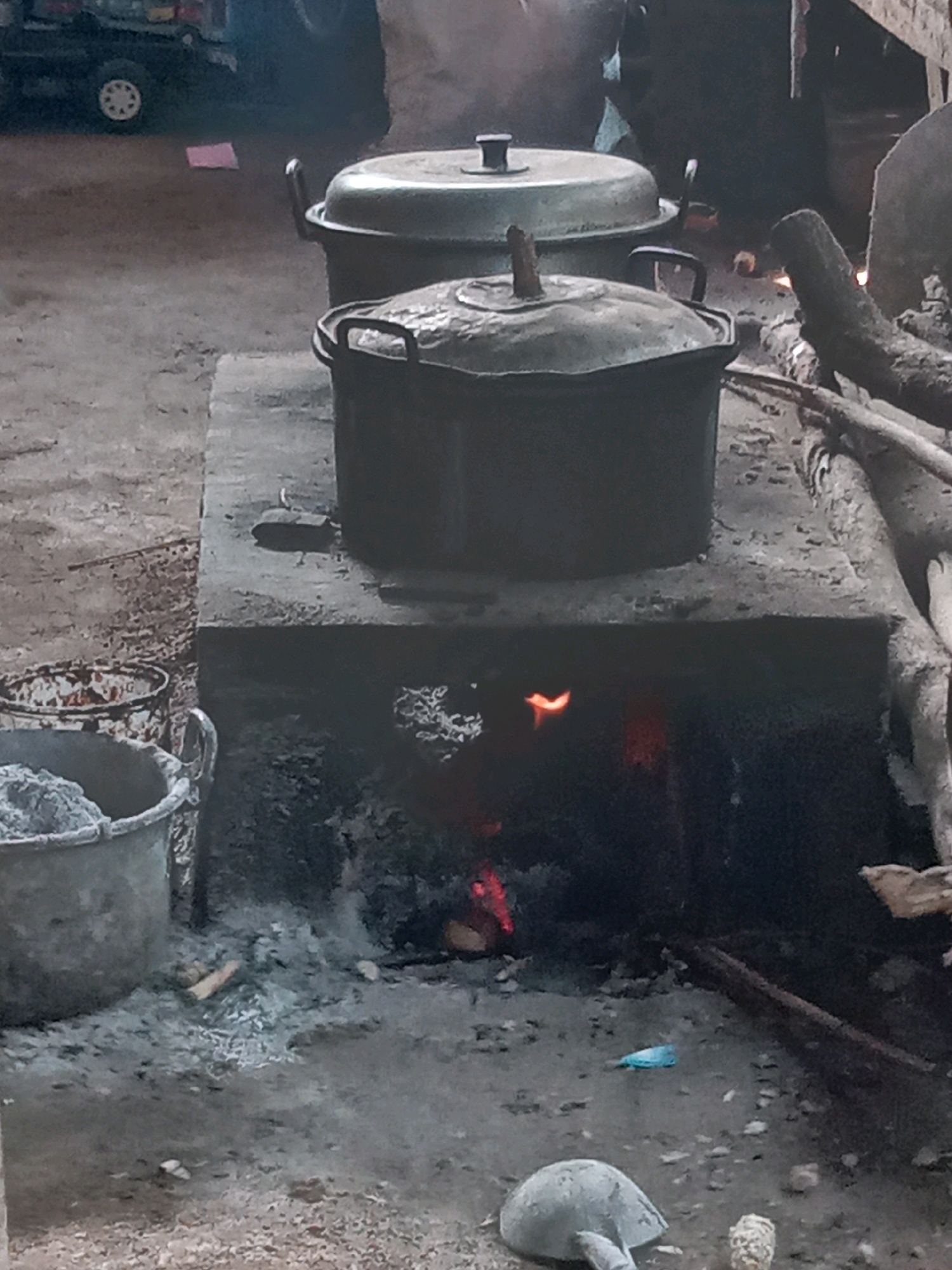 A medium shot captures two large, dark cooking pots placed on top of a makeshift concrete stove in Sukoanyar, Indonesia. The stove has a rectangular opening at the bottom, through which a fire is visible, casting a reddish glow. Ashes and embers surround the opening. The larger pot in the foreground has a slightly dented lid with a wooden handle. The pot behind it, slightly elevated, also has a lid with a handle. To the left of the stove, two large, grey buckets are visible. One bucket is partially filled with a grey granular substance, possibly cement or sand, and the other, to its left, appears empty and stained. The ground surrounding the stove and buckets is rough and uneven, with scattered debris, including a small blue plastic item. In the background, a blurred image of a truck with a white wheel is present, along with a pink rectangular object on the ground. The overall scene suggests an outdoor cooking or construction setting.