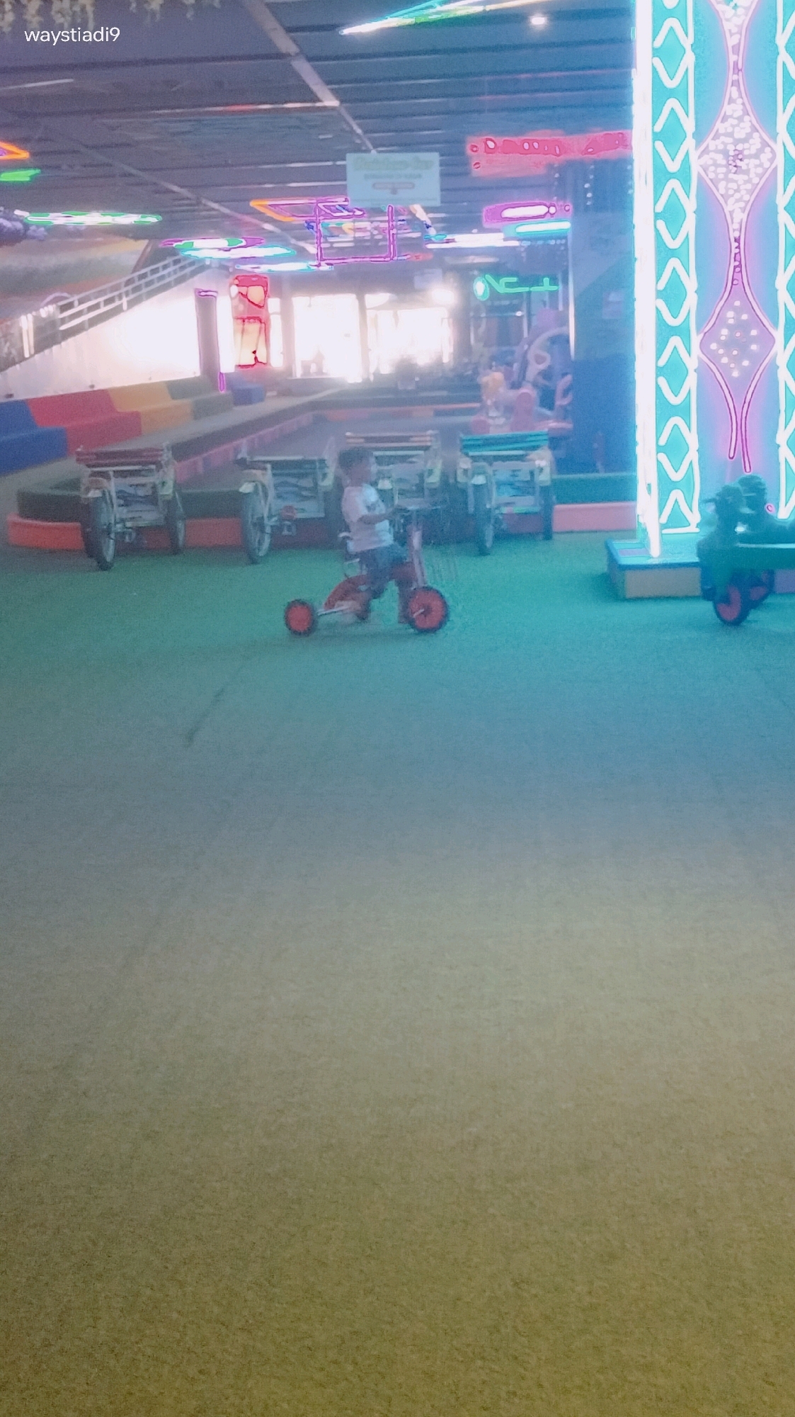 The image depicts an indoor amusement area, possibly an arcade or entertainment center, with a young child riding a red tricycle in the foreground. The child is wearing a light-colored t-shirt and dark shorts. Behind the child, there are several stationary ride vehicles, some of which appear to be miniature versions of cars or trucks. The background features brightly lit neon signs and decorative elements, including a tall, vertical, illuminated structure with a diamond pattern. The floor is covered in a green carpet-like material. The overall impression is a colorful and lively environment designed for entertainment. The text "waystiadi9" is overlaid in the top left corner of the image. Without further context, it is not possible to definitively pinpoint the specific location within Bandung City, Indonesia.