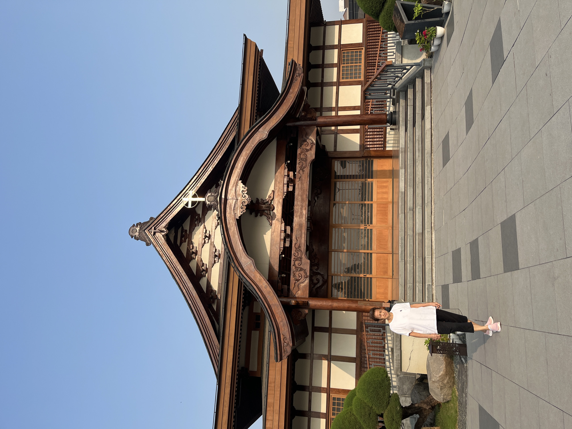 An elderly Asian woman with short, dark hair is standing on a paved walkway in front of a large, traditional-style building with a wooden exterior and a multi-tiered, curved roof. She is wearing a white t-shirt and dark pants, with pink athletic shoes. The building features intricate wooden carvings, large wooden doors, and ornate window frames. A stylized cross is visible at the apex of the roof, suggesting it might be a religious institution. To the left of the building, there are manicured bushes and large rocks. To the right, there are stairs leading up to the building's entrance, with potted plants along the railing. The sky above is clear and blue. The paving stones are large and square, with some darker rectangular insets.
