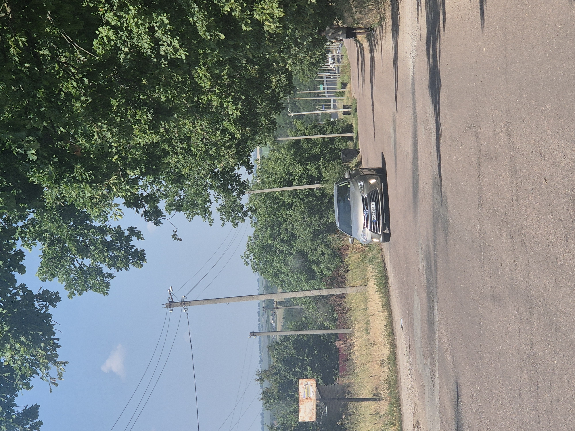 A silver sedan is driving down a paved road in Pidhaitsi, Ukraine, on a clear day. The road is lined with trees and utility poles. A person is jogging on the right side of the road. In the background, there are fields and more trees. The car's license plate is partially visible and reads "BA 3350 BX".