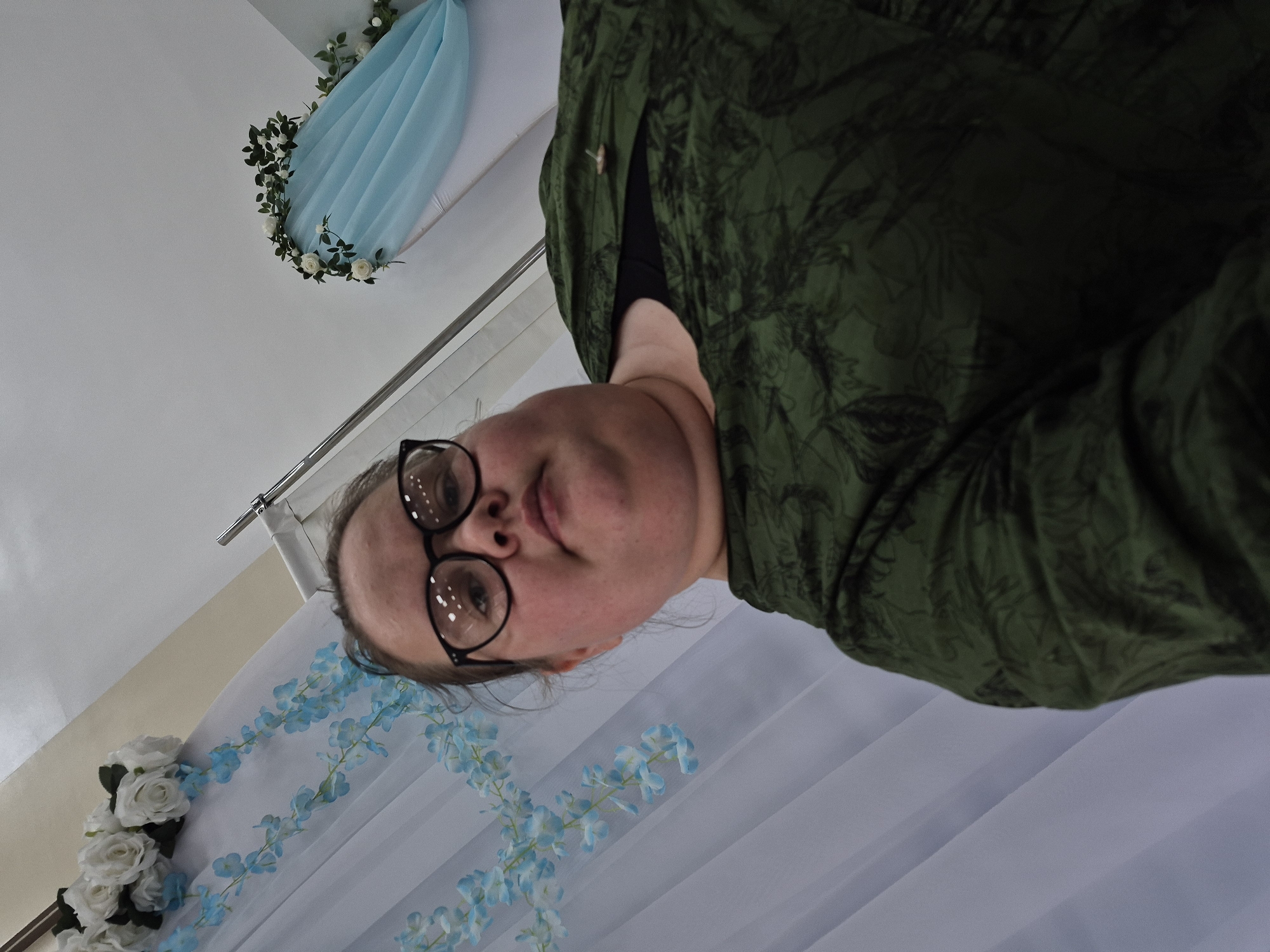 An upward-facing, rotated portrait captures an adult female wearing black-rimmed glasses and a dark green top with a subtle, leafy pattern. Her face, seen from a low angle, is centered on the right side of the frame, with her gaze directed towards the camera. The background suggests an indoor setting, likely a decorated venue in Chaikovsky, Russia.
To the upper left, a light blue fabric is elegantly draped and adorned with a garland of artificial white roses and green foliage. Below the subject's face, a sheer white curtain features a delicate light blue floral pattern, possibly of wisteria, suggesting a decorative backdrop for an event. A metal curtain rod is visible near the top of the background. The lighting is soft and diffused, characteristic of an indoor environment, with no explicit indicators of the time of day or external weather conditions. No text is visible in the image. The composition implies either a selfie or a photograph taken from a very low viewpoint, potentially with the subject leaning down towards the camera.
