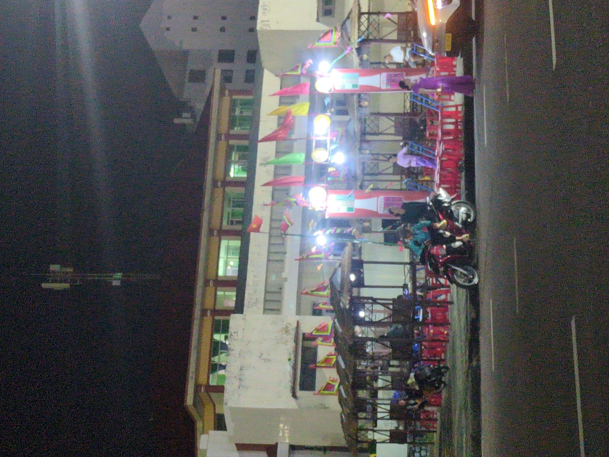 The scene depicts an evening in Tuy Hòa, Vietnam, with a brightly lit multi-story building adorned with numerous small, colorful flags vertically. The building is likely a restaurant or commercial establishment due to the presence of tables and chairs visible on its levels. People are present on the building's levels, some wearing traditional Ao Dai dresses. Several motorcycles are parked on the street beside the building. The night sky is dark, with faint light sources in the distance, possibly indicating other buildings or infrastructure. A communication tower is seen silhouetted against the dark sky. The road surface appears to be asphalt with painted lane markings.