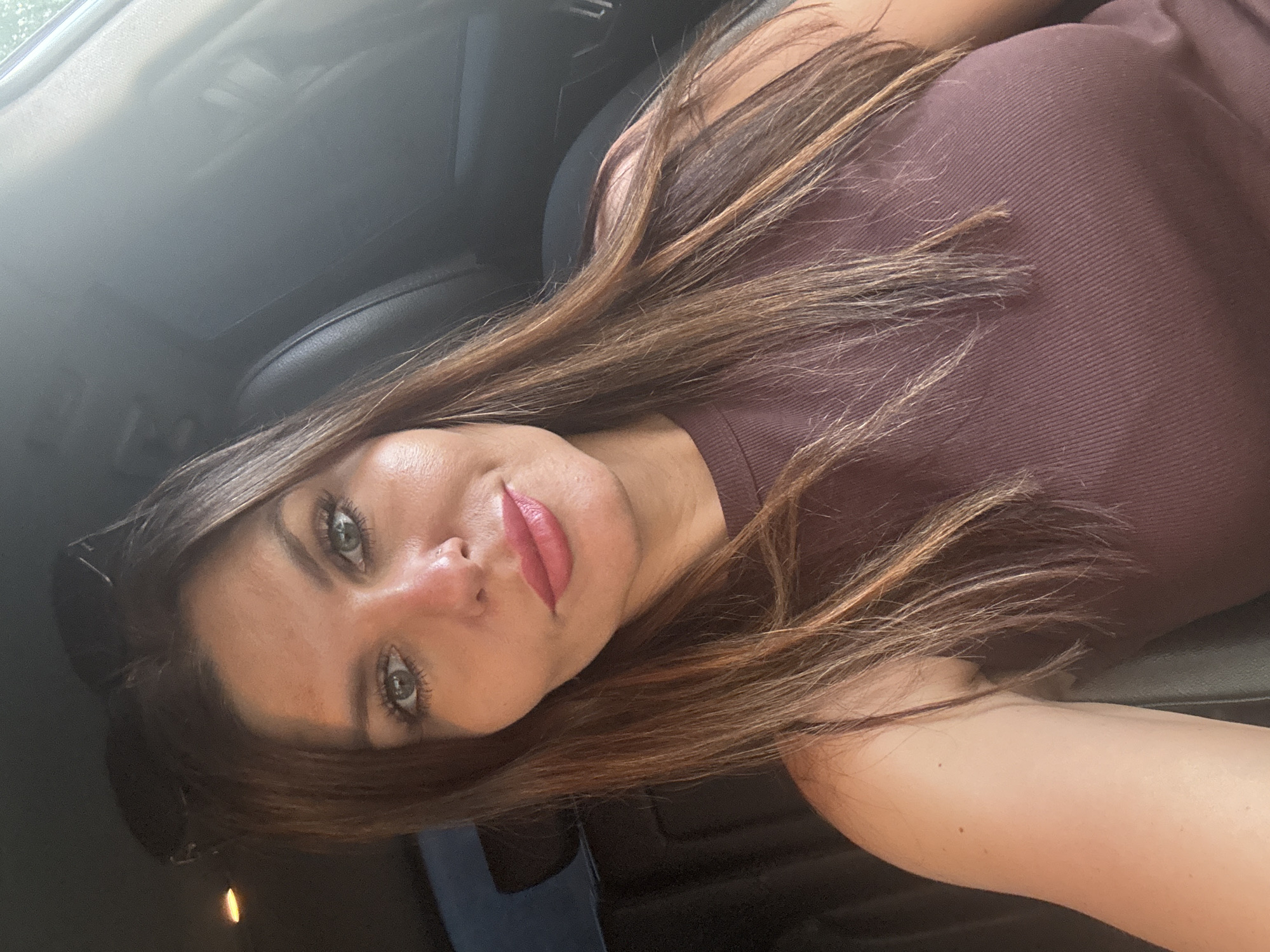 A woman with long brown hair and green eyes is taking a selfie. She is wearing a brown top and has pink lipstick on. She is inside a car. The location is not discernible.
