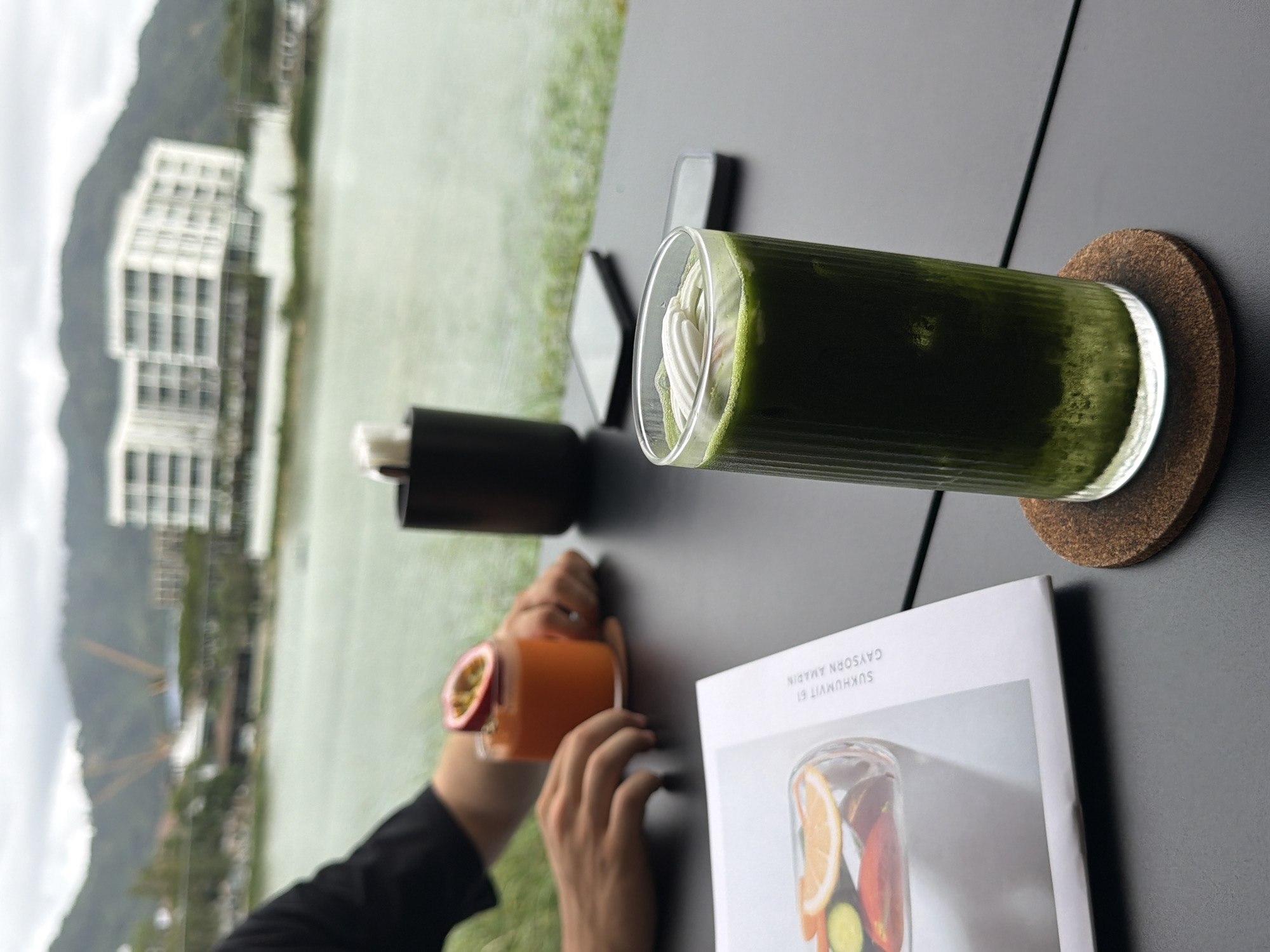 The image shows a table setting with drinks and a magazine, likely outdoors in Choeng Thale, Thailand. A tall glass of a dark green liquid with a swirl of white cream on top sits on a circular cork coaster on a dark gray table. A second drink, light orange in color and topped with a halved passion fruit, is held by a person's hands. A black cylindrical container, likely for napkins or utensils, is also on the table. A white magazine with an image of a fruit-filled jar on its cover partially sits near the passion fruit drink. The cover is legible: "Gaysorn Amarin, Sukhumvit 61." In the background, blurred out, is what appears to be a body of water, possibly a lake or bay, with a modern white building with many windows on the opposite shore. Hills or mountains are also visible in the distance.