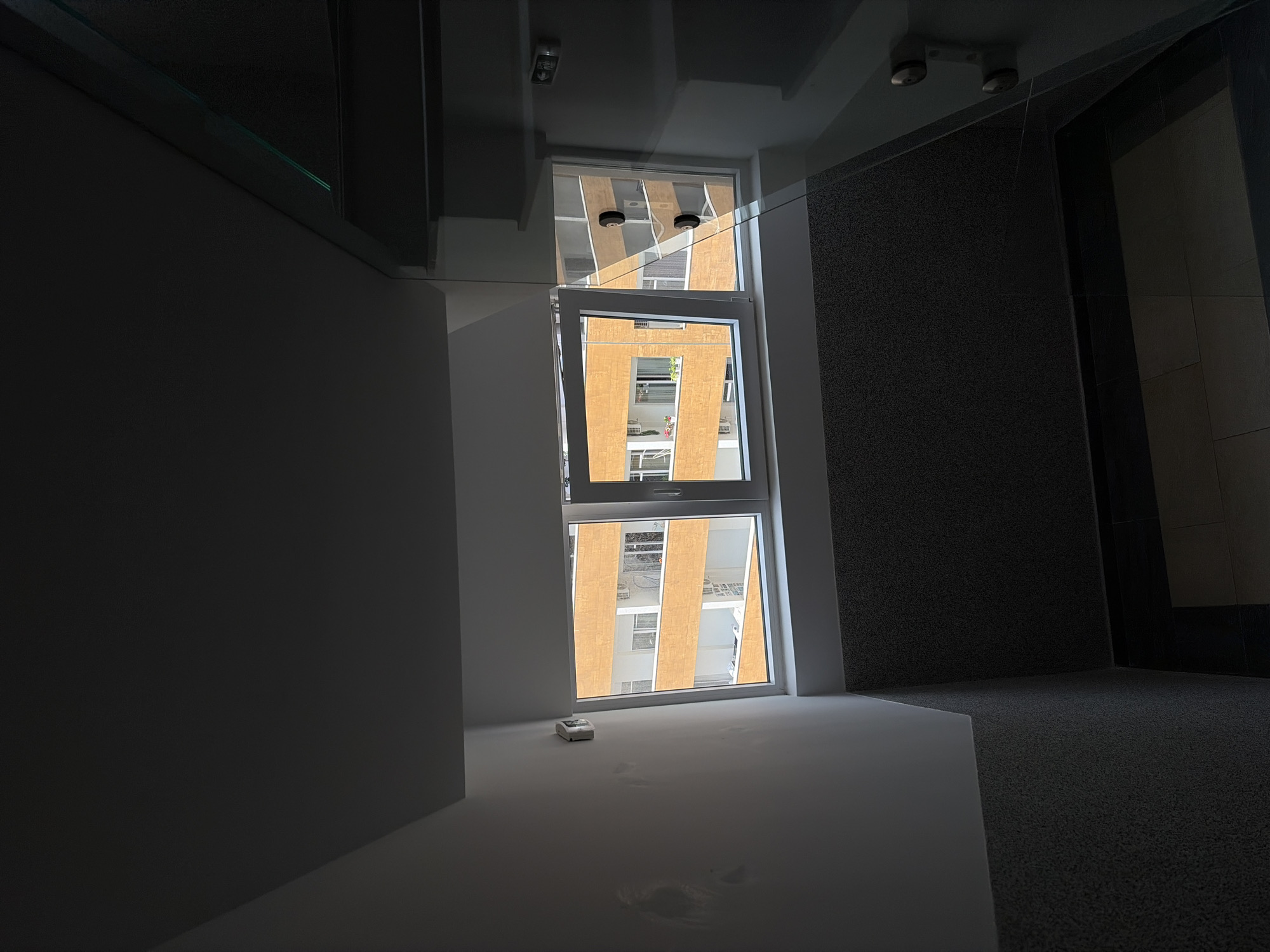 The image depicts an interior space with a large, multi-section window looking out onto a building with a distinctive facade. The view through the window shows a building with a repeating pattern of orange-colored vertical stripes and white balconies.
The interior space has a modern aesthetic with clean lines and predominantly gray and white color scheme. There's a stark contrast in lighting, with bright light streaming through the window and casting shadows on the white surfaces. The presence of footprints on the white surface implies human activity in the space. A small, rectangular object, possibly a sensor or device, is located near the window.
Based on the building visible through the window and its architectural style, the location is consistent with contemporary developments in Budva, Montenegro. However, without additional specific landmarks, it is impossible to pinpoint the exact location within the city.