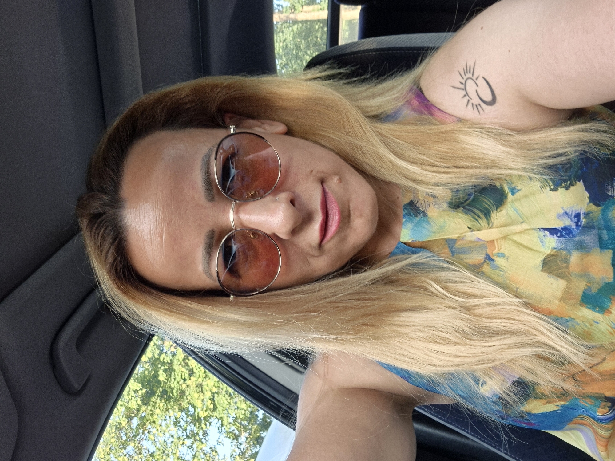 A medium shot shows a woman in sunglasses and a colorful dress, sitting in the driver's seat of a car. Her blonde hair is down and reaches her shoulders. She is wearing round, rose-tinted sunglasses and has a faint smile. A tattoo of a crescent moon with sun rays is visible on her left shoulder. The car's interior is dark, with a glimpse of greenery and trees through the open window on the left. The background is blurred, suggesting the car is in motion.