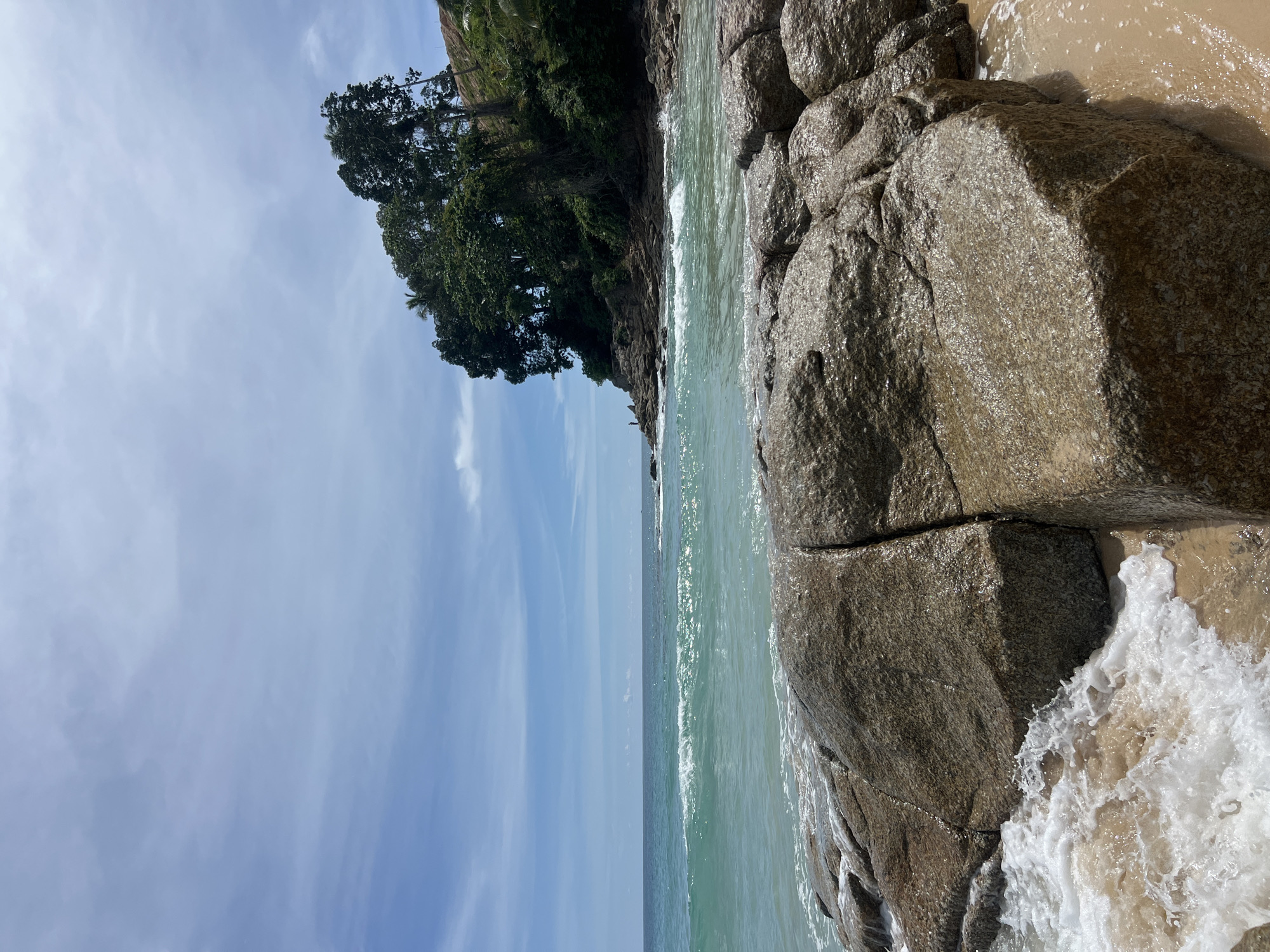 This image depicts a rocky beach scene on the coast of Choeng Thale, Thailand. The foreground is dominated by large, wet, tan-colored rocks with visible cracks, partially covered by frothy white waves and light brown sand. The ocean water is a clear turquoise with white-capped waves breaking closer to the shore. In the midground, the water stretches to the horizon under a blue sky with scattered white clouds. To the right, a small, dark-haired figure can be seen standing on a rocky outcrop. Beyond the water and outcrop, a lush green hillside covered in dense trees rises, with more scattered clouds in the sky above. The overall impression is a natural, tropical coastal environment.