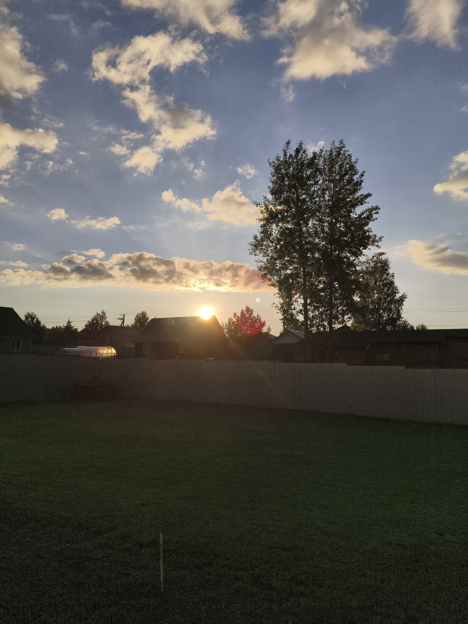 This media file presents a peaceful rural or suburban landscape at sunset, located in Русский Вожой, Russia. The image is composed of distinct horizontal layers, showcasing the transition from a shadowed foreground to a brightly lit sky.
In the foreground, a wide stretch of dark green grass covers the lower third of the frame, appearing almost black due to the low sun angle and deep shadows. A small, light-colored stick or marker is subtly visible in the grass near the bottom center.
A long, light-colored fence, possibly made of wood or metal, spans horizontally across the midground, separating the grassy area from the properties beyond. Behind the fence, a collection of residential houses with dark roofs and a dense array of trees are silhouetted against the vibrant sky. The sun, a brilliant golden orb, is positioned low on the horizon, partially obscured by the houses and trees. Its intense light creates a prominent sunburst effect, backlighting the landscape and casting most elements into deep shadow. A notable feature among the houses is a greenhouse or poly-tunnel, which brightly reflects the sun's warm glow. A tall, leafy tree stands prominently on the right side of the frame, with other trees of varying heights forming a canopy behind the houses.
The sky in the upper portion of the image displays a gradient from soft blue at the top to warmer hues closer to the horizon. It is dotted with scattered cumulus clouds, which catch the golden and orange light of the setting sun, particularly those closer to the horizon. The overall lighting indicates either late afternoon or early evening.
The scene is devoid of people or specific activities, creating a serene and tranquil atmosphere that emphasizes the natural beauty of the sunset over a quiet residential area. No visible text is present in the image.