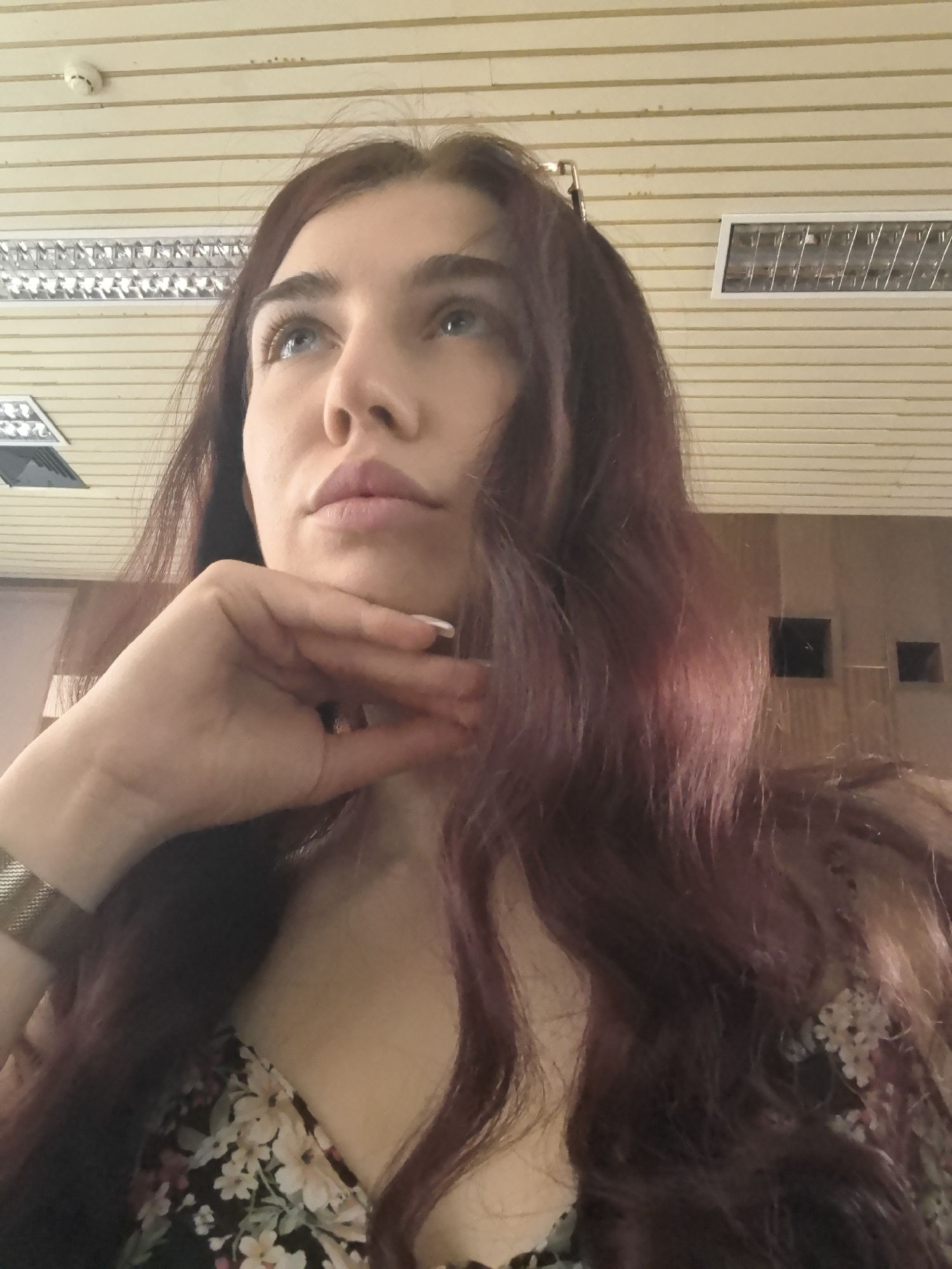 A medium shot captures a woman with dark auburn hair looking upwards, her chin resting on her left hand. She is wearing a floral print top. The background appears to be an indoor setting with a slatted ceiling and fluorescent lighting. The image is taken from a low angle, looking up at the woman. There are no visible landmarks or features that specifically place the image in Stara Zagora, Bulgaria.