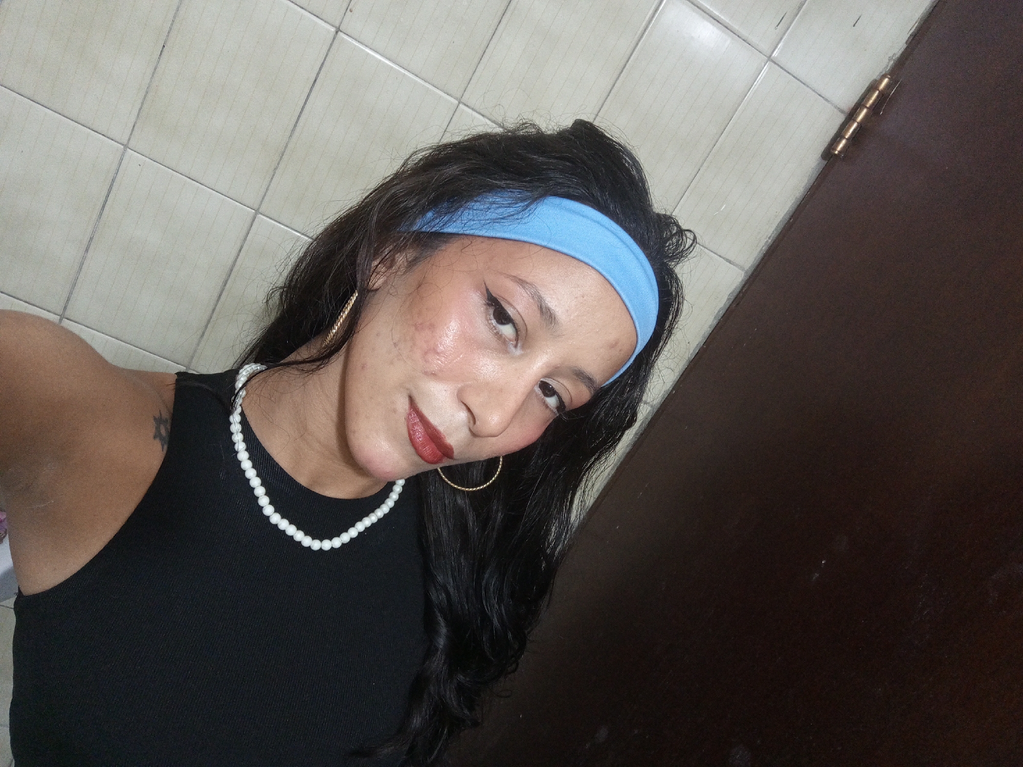 A woman is taking a selfie. She is wearing a black tank top, a pearl necklace, and a blue headband. She has gold hoop earrings and dark lipstick. The background includes a tiled wall and a dark brown door with a gold hinge. The photograph's location and city of Porlamar, Venezuela, cannot be determined from the available image information.