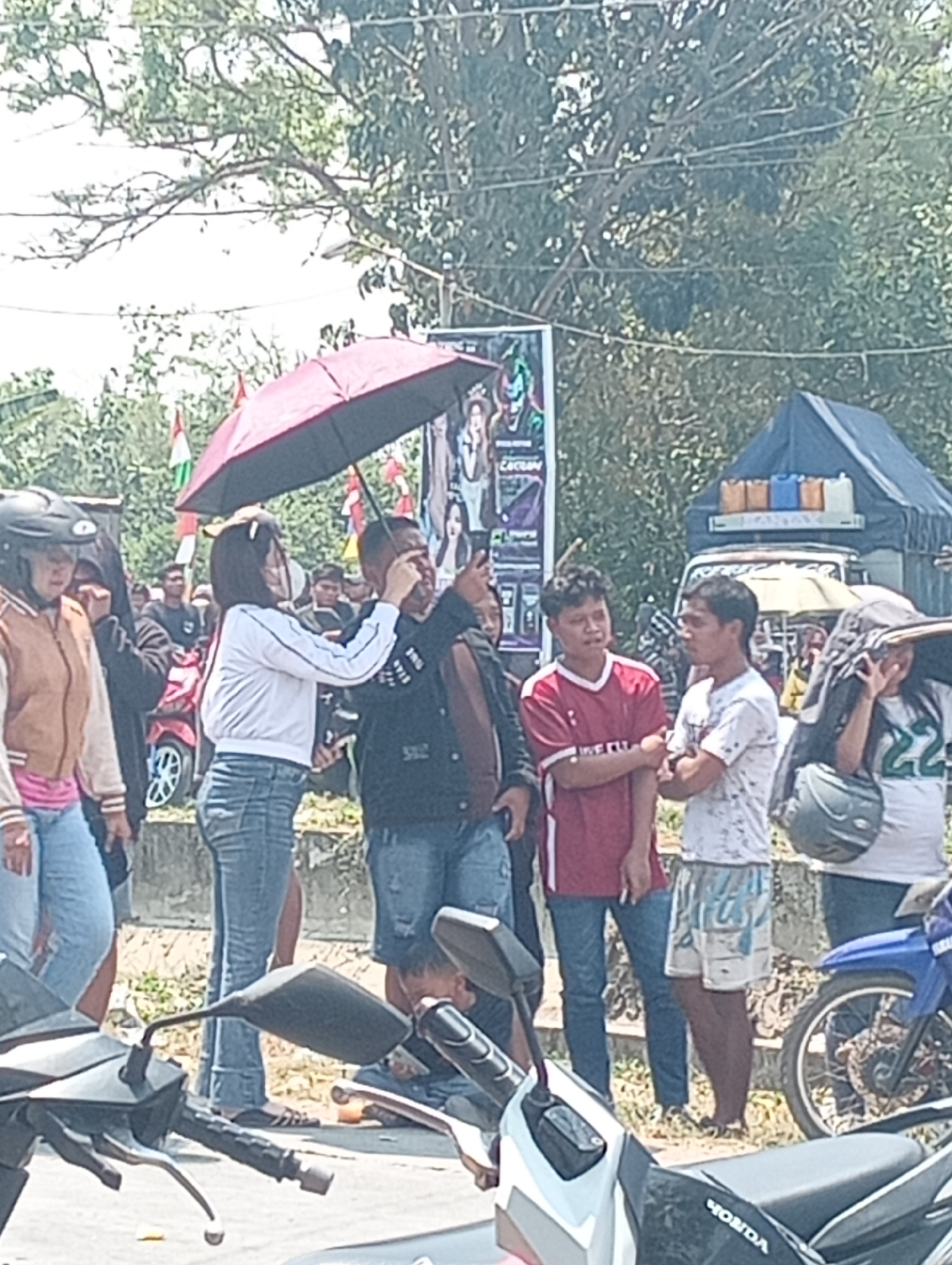 A group of people are gathered outdoors, with a focus on a woman holding a red umbrella over herself and another person. She is wearing a white shirt and blue jeans, and the other person is wearing a dark jacket and jeans. The scene appears to be in a public area, possibly a street or market, with trees and a tent visible in the background. Several motorcycles are parked in the foreground. The overall impression is that of a casual outdoor gathering in a warm climate.