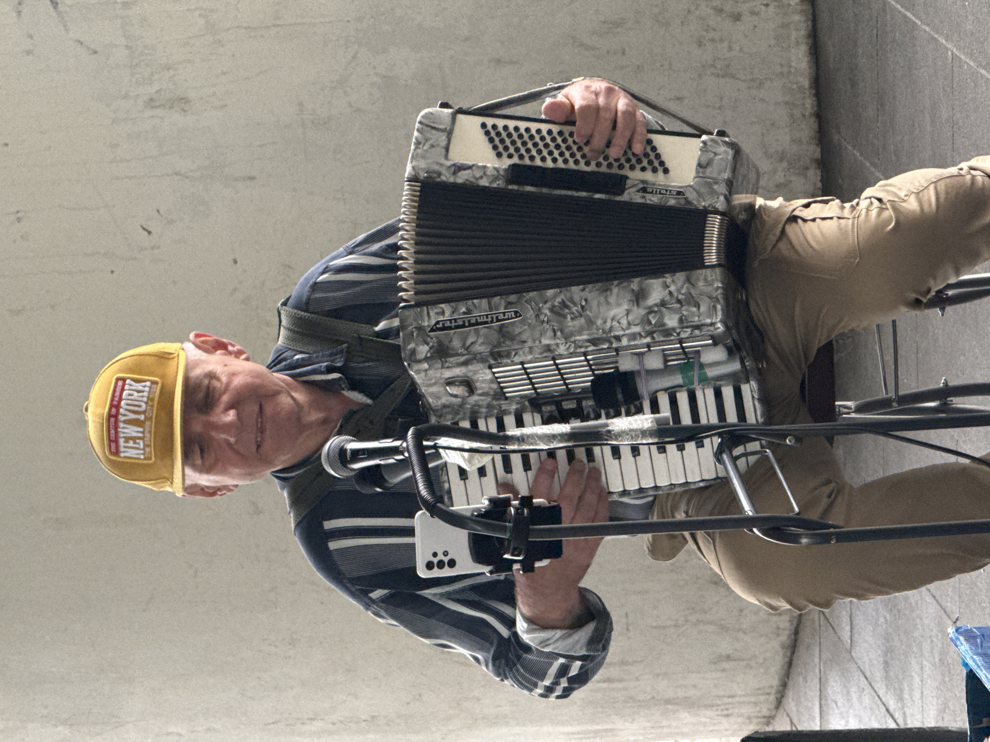 An elderly man wearing a yellow "New York" baseball cap and a striped blue and white shirt plays an accordion. He is seated on a stool in front of a microphone stand with a mobile phone attached. The accordion has a marbled grey and white casing with black bellows. The man's hands are on the accordion keys and bellows. He is wearing khaki pants. The background appears to be a grey concrete wall and a pillar. The man is looking down and appears to be singing into the microphone. The scene is lit by natural light. It is not possible to determine the location in the city of Moscow, Russia from the image.