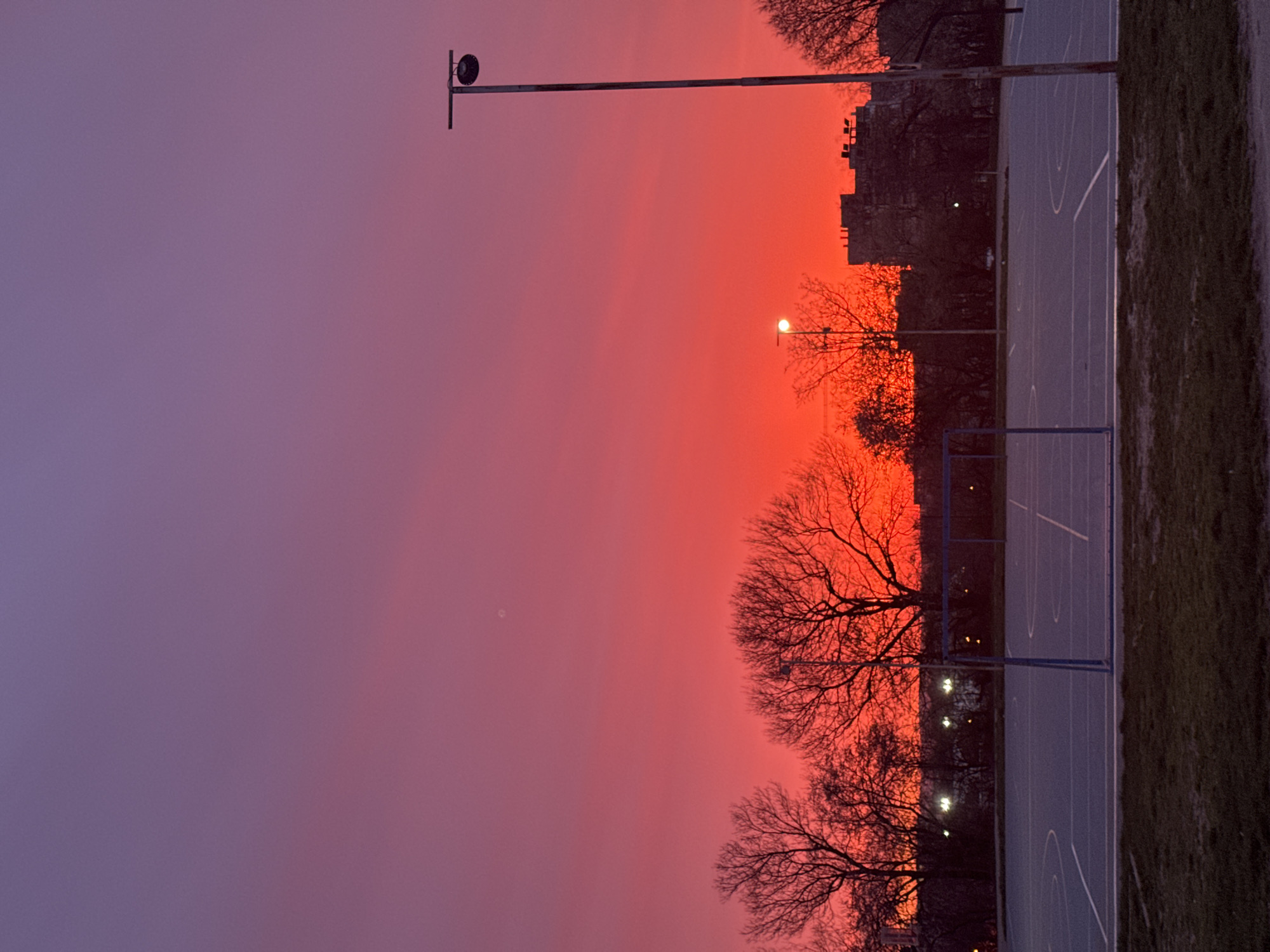The image depicts a basketball court located in Varna, Bulgaria, during a vibrant sunset. The sky transitions from a deep purple on the left to a fiery orange-red on the right, indicating the setting sun. Silhouetted against the sky are several bare trees, a building, and lampposts. One lamppost is directly in front of the court while others can be seen in the background. The court itself is painted blue with white lines marking the boundaries and arcs. A basketball hoop and backboard are visible at one end of the court. A patch of grass borders the right side of the court.