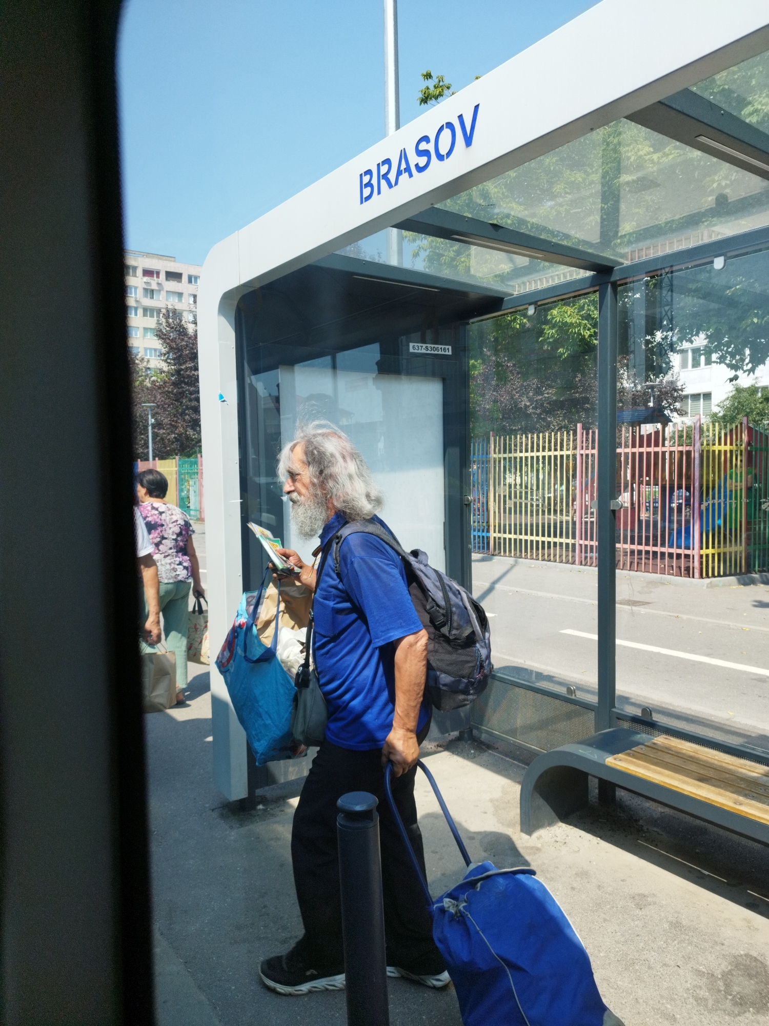 The image shows a man with long gray hair and a beard standing at a bus stop. He is wearing a blue t-shirt and black pants, and is holding a book and a blue bag. He is also pulling a blue wheeled bag behind him. In the background, there is a building and a colorful fence. The bus stop has the word "BRASOV" written on it. The image is taken during the day, and the sky is clear and blue. The scene appears to be a street in the city of Bucharest, Romania.