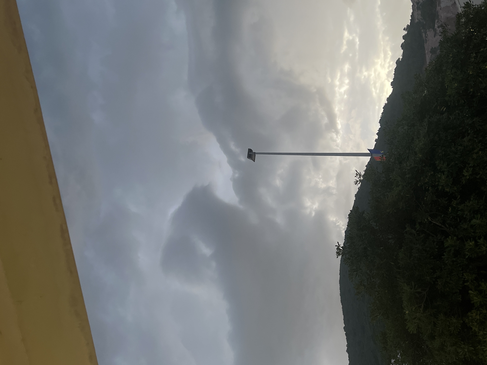 The image, taken outdoors in overcast conditions, shows a gray sky, possibly in Budva, Montenegro. A mountain covered with dense green vegetation is seen in the background, to the right. A tall pole stands in the middle, possibly a flagpole. Part of a yellow building is visible on the left edge of the frame.