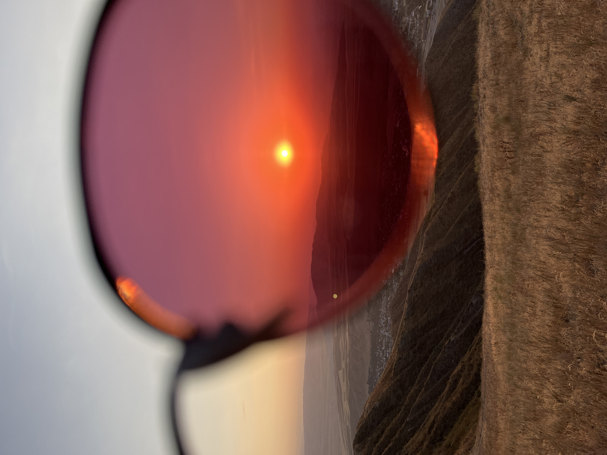 A close-up, eye-level shot captures a sunset as viewed through pink-tinted sunglasses. The sunglasses frame the upper portion of the image, their lenses reflecting a brilliant orange sun low on the horizon, casting a warm glow over distant, silhouetted hills. In the foreground, the muted tones of dry, grassy hills descend towards a small town visible in the middle ground, nestled within a valley. The sky transitions from a pale blue at the top to a soft yellow-orange near the horizon.