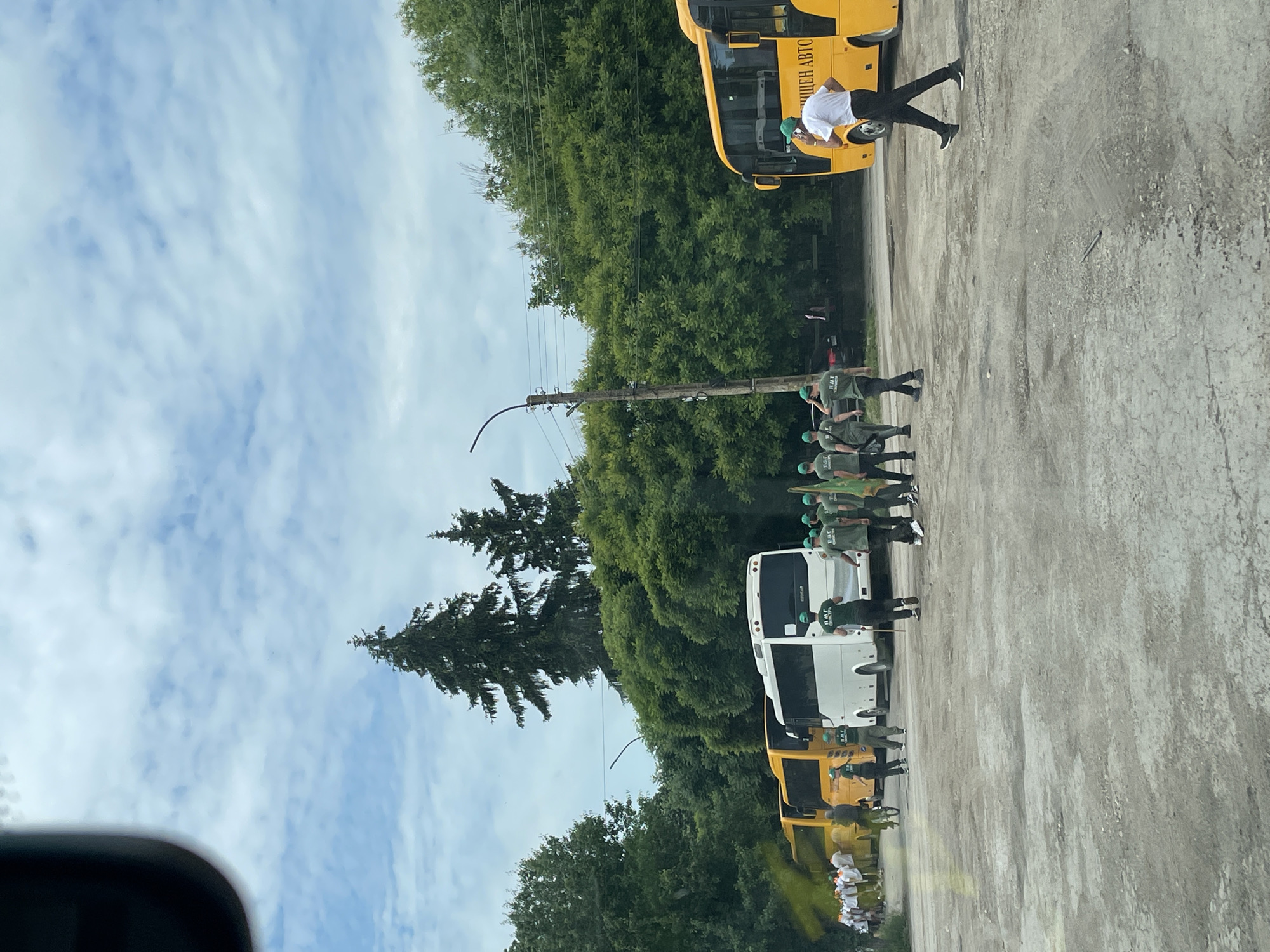 The scene is an outdoor area, likely a parking lot or a gathering space, in Tsar Kaloyan, Bulgaria. The sky is partly cloudy. The ground is concrete. A line of people in dark green uniforms and light green caps stands in formation. Some are holding green flags or poles. In the background are buses, some are yellow, some are white. One man is standing by a yellow bus wearing a white shirt and dark pants. A line of trees runs across the center of the image. A utility pole stands among the trees.