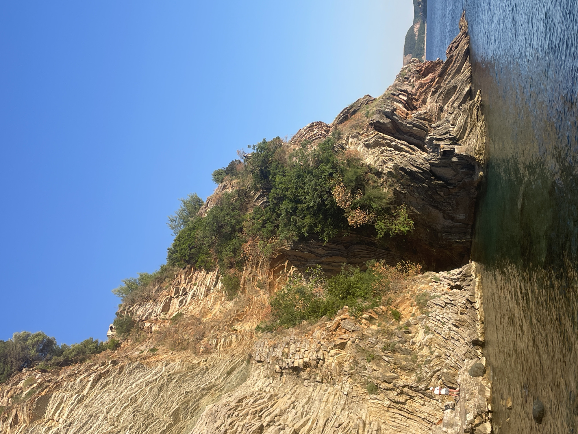 The image shows a rocky cliff face covered in vegetation in Rafailovići, Montenegro, next to the sea. The rocks are layered and appear to be sedimentary. Two figures are lying on the rocks near the water. The sea is calm. The sky is clear and blue.