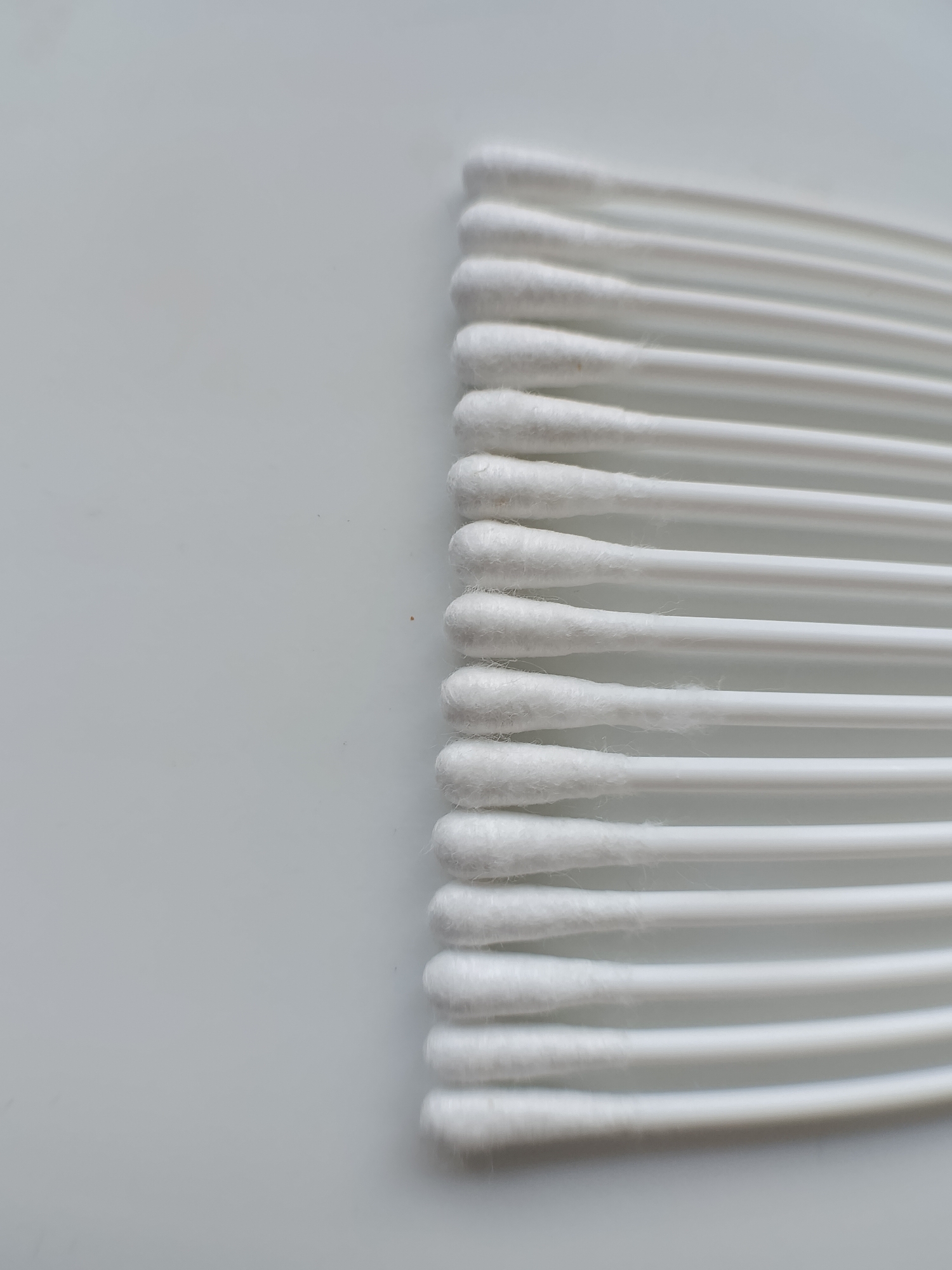 A close-up, high-angle shot displays a row of white cotton swabs arranged horizontally on a plain white background. The swabs are positioned on the right side of the frame, with their cotton tips facing upwards and to the left. The background is a smooth, seamless surface, creating a minimalist and sterile environment. The lighting is even and diffused, highlighting the texture of the cotton tips and the smooth surface of the plastic handles. There are no discernible environmental features or contextual elements that would suggest a specific city or location.