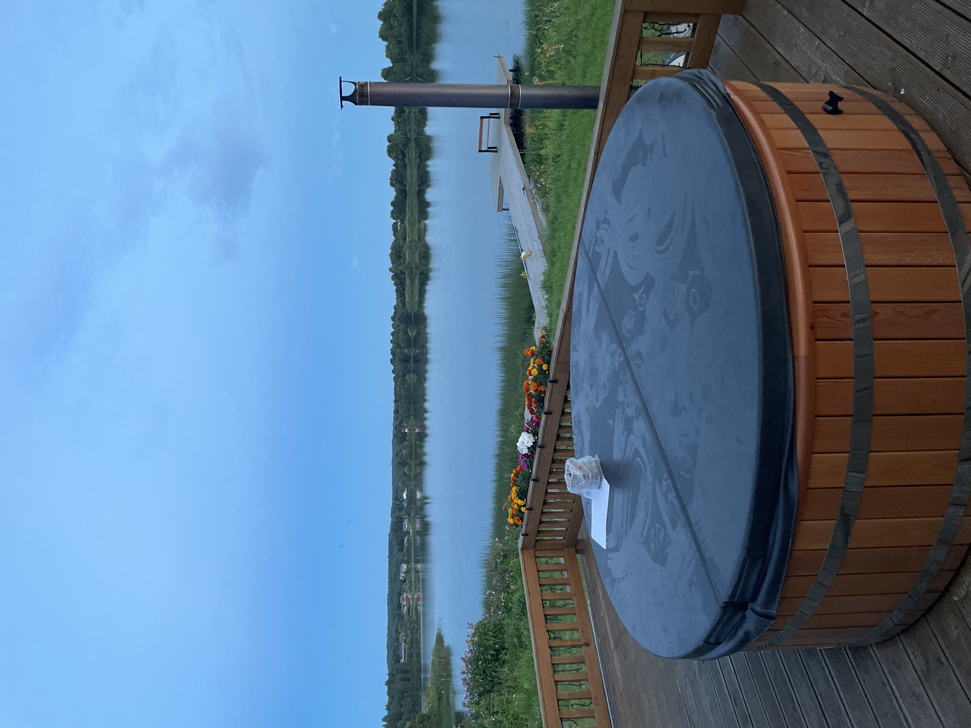 A round wooden hot tub covered with a dark gray lid sits on a wooden deck. The deck is bordered by a wooden railing with flower boxes filled with colorful flowers. Beyond the deck is a large body of water, likely a lake, reflecting the overcast blue sky. A wooden pier with a bench extends into the water. In the background, a treeline with houses and a chimney is visible. The scene appears to be set in the city of Pyhäjärvi, Russia.