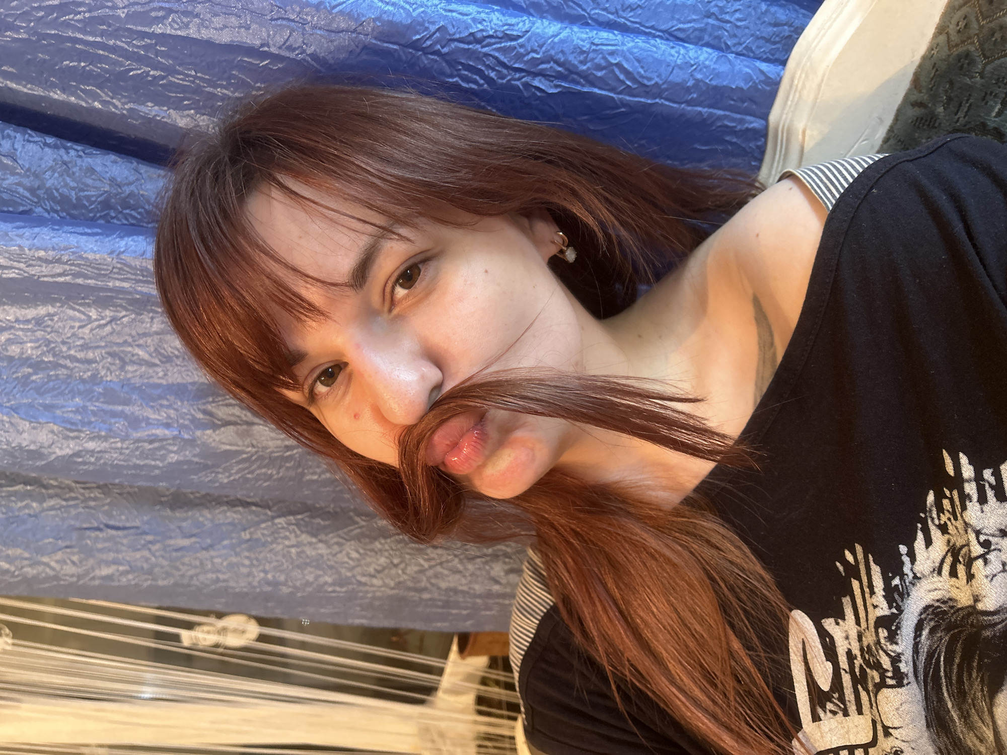 A young adult female is posing for a photograph. She is lying down with a blue, crinkled fabric behind her. She is wearing a black t-shirt with a white graphic. The woman has auburn hair, with strands held under her nose to mimic a mustache. She is looking directly at the camera. The location is not discernible from the image.
