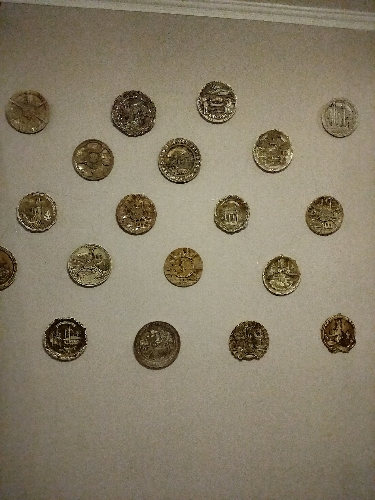 The image shows a collection of approximately 20 circular metal objects, resembling coins or medals, displayed on a lightly textured, beige wall. The objects are of varying sizes and feature intricate embossed designs. Some coins depict architectural structures, such as buildings and columns, while others show scenes with figures and possibly landscapes or ships. The overall lighting suggests an indoor setting, potentially a museum or exhibition, as the objects are mounted on the wall in a deliberate arrangement. There are no people visible in the image. The wall has a slight crown molding at the top. There is no visible indication of the city of Nizhnedonskoy, Russia.