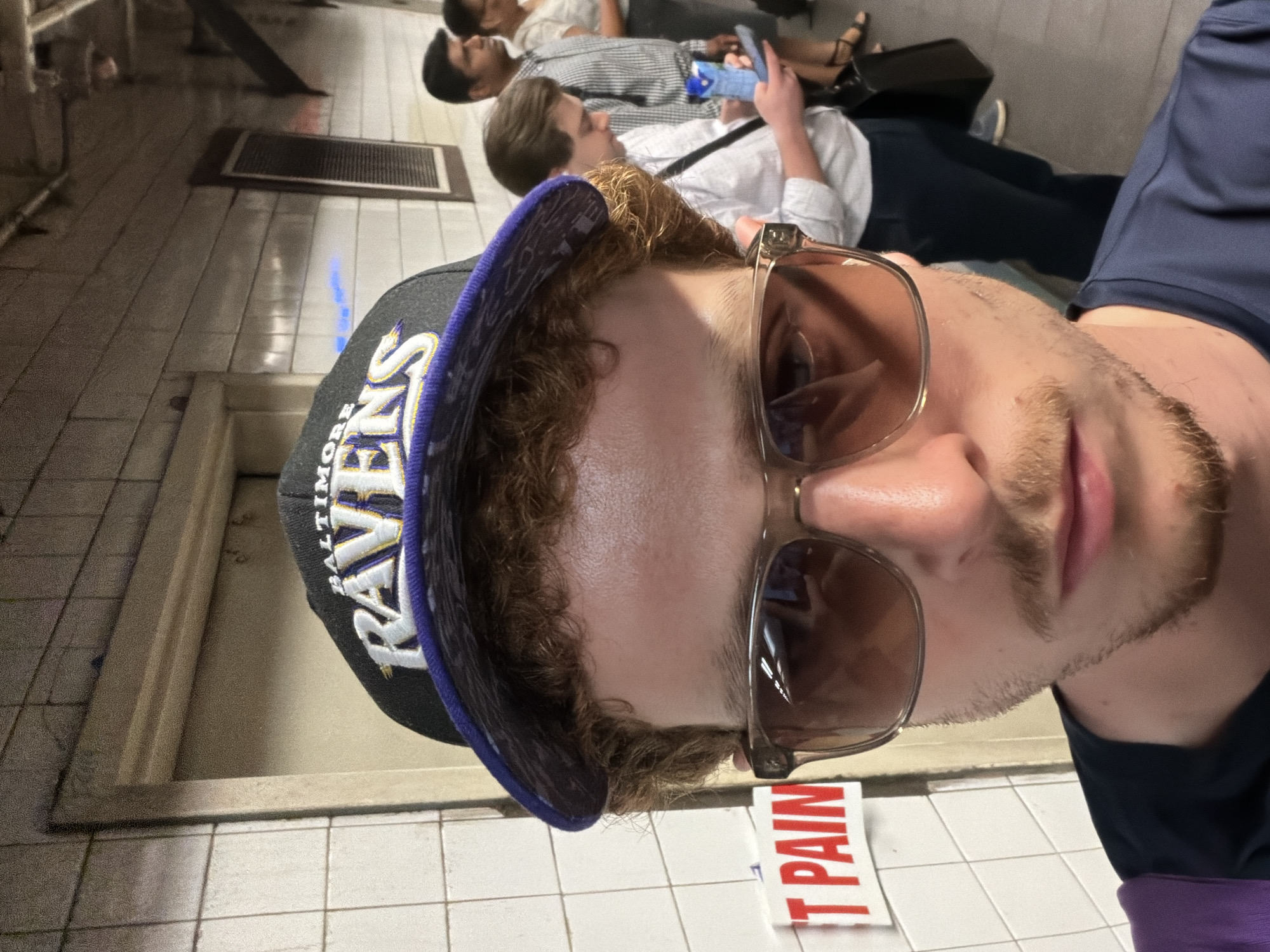 A medium close-up photograph shows a person wearing a Baltimore Ravens baseball cap and tinted sunglasses, standing on a subway platform. Behind the person, other individuals are seen sitting on a bench and standing. The subway platform is tiled with white square tiles. A sign reading "IT PAIN" is visible at the bottom of the frame. The location is likely a subway station in the city of New York, United States of America.