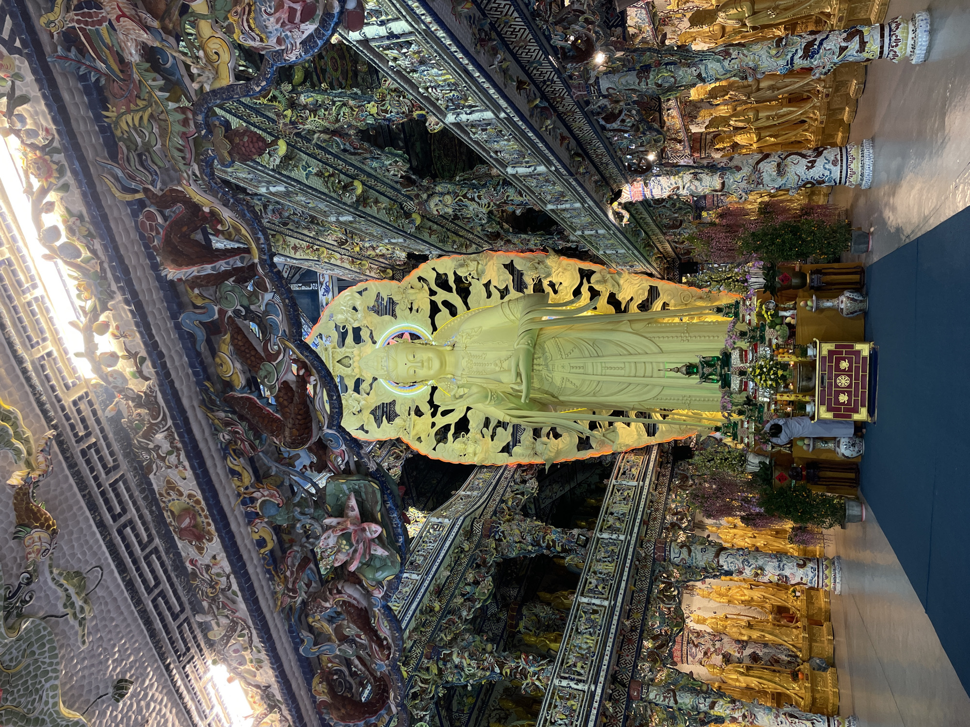 The image shows a view inside the Bửu Long Pagoda in Xuân Thành, Vietnam. A large, golden statue of a standing Buddha dominates the central space. The interior architecture and decor are intricately designed and decorated, featuring colorful carvings, mirrored surfaces, and detailed mosaics. Multiple golden statues and columns are situated along the perimeter of the prayer hall. A person dressed in white is praying. Potted plants and ceremonial items are visible near the statues.