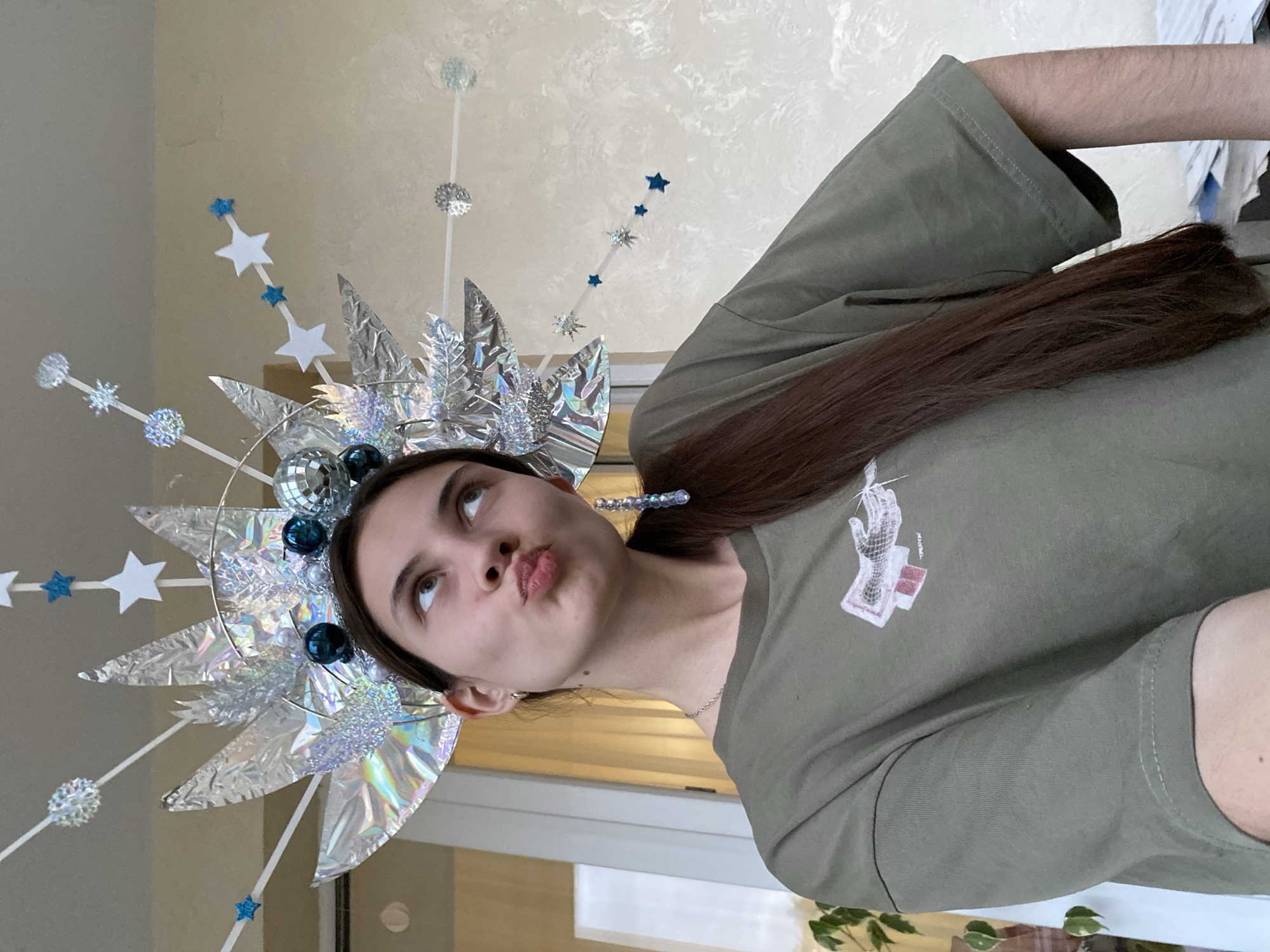 A young woman with long brown hair is posing for a photo while wearing an elaborate, iridescent headdress. The headdress is adorned with silver foil spikes, blue and silver stars, snowflakes, and small disco balls. She has a playful expression, with her lips pursed and eyes looking upwards. She is wearing a green t-shirt with a graphic print of a hand holding a document. The background appears to be indoors, with a wall and a window visible. There is no indication of the location being Votkinsk, Russia.