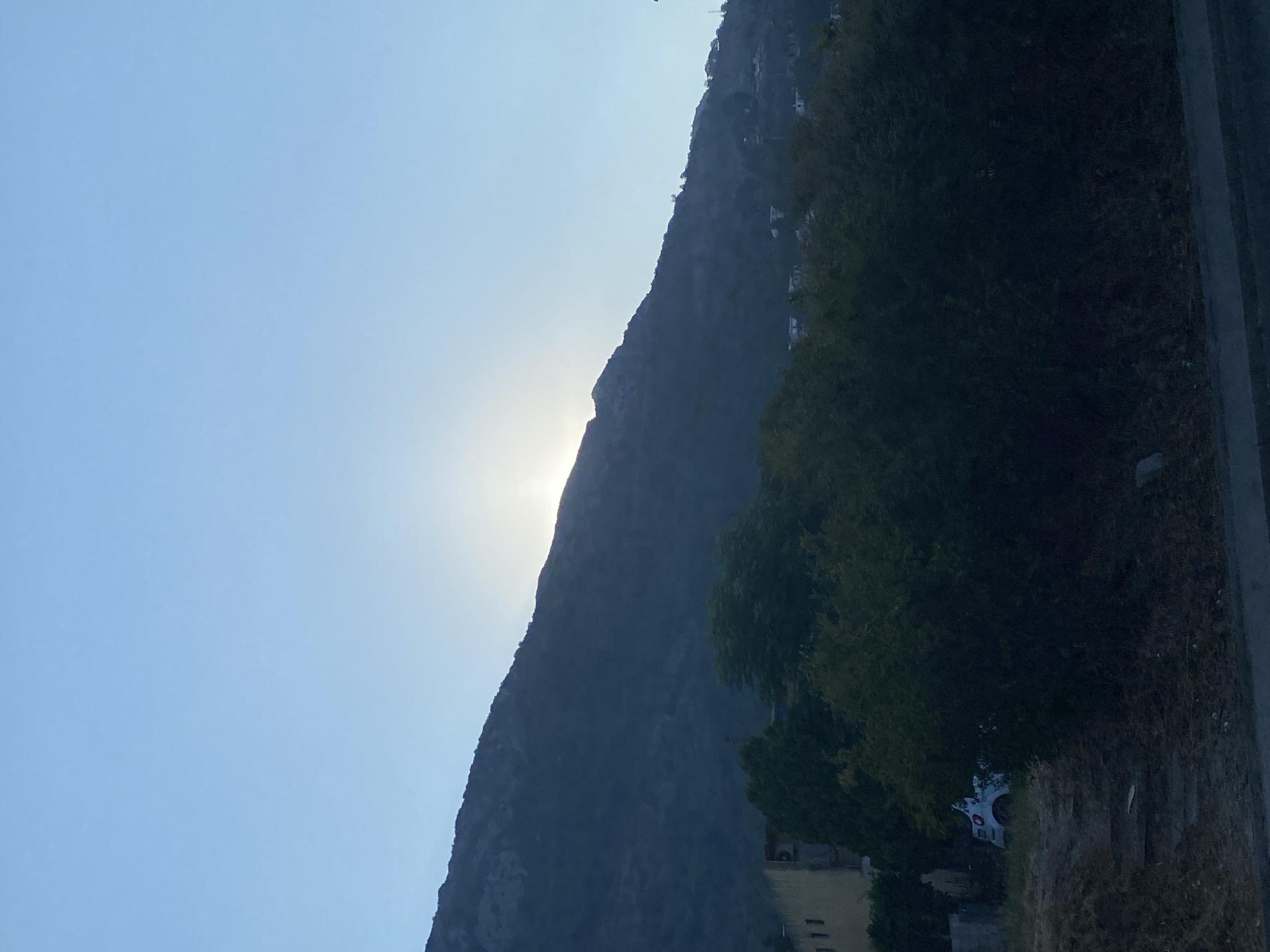 The image captures a serene landscape with a prominent mountain range in the background, partially obscured by the rising or setting sun, casting a soft glow over the scene. The sky is a gradient of pale blue, transitioning to a deeper hue near the horizon. In the mid-ground, dense foliage of trees and shrubs creates a natural barrier. To the left, a partially visible building with windows can be seen, suggesting a human settlement. Further left, a small white vehicle is parked amidst the greenery. The foreground consists of dry grass and some scattered debris, with a concrete structure or road edge at the bottom. The overall impression is of a quiet, natural setting on the outskirts of a town, likely Sutomore, Montenegro, as indicated by its characteristic hilly terrain and proximity to vegetation. There are no people or observable actions in the image.