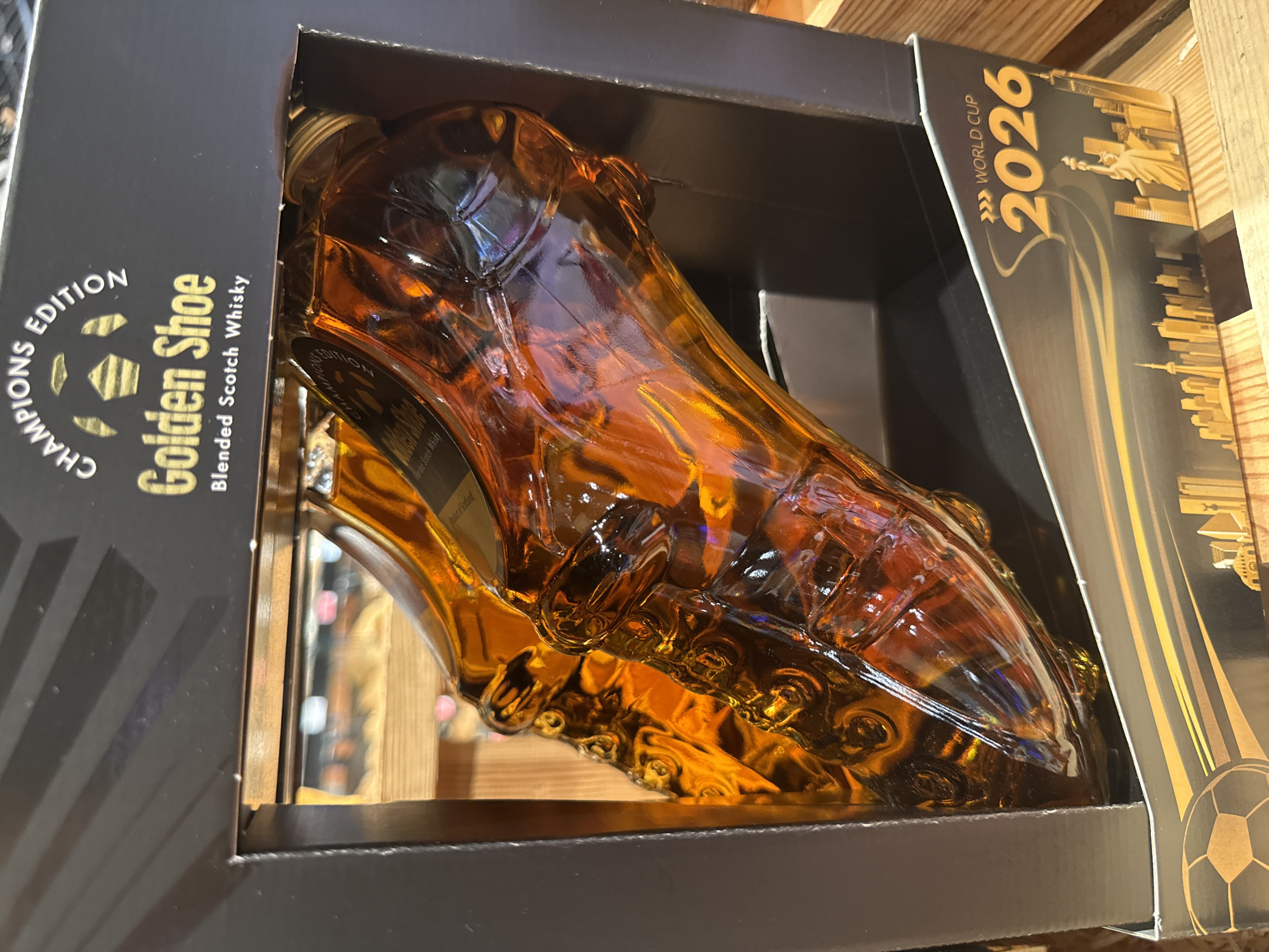 The image shows a gift set of "Golden Shoe" blended Scotch whisky, packaged in a black box with a soccer-themed design. The bottles are shaped like soccer cleats and are filled with amber-colored liquid. The box features the text "CHAMPIONS EDITION" and "WORLD CUP 2026" along with an illustration of a city skyline, including the Statue of Liberty. The product is displayed on a wooden shelf. The photograph was taken at a store in Herford, Germany, on September 3, 2023.