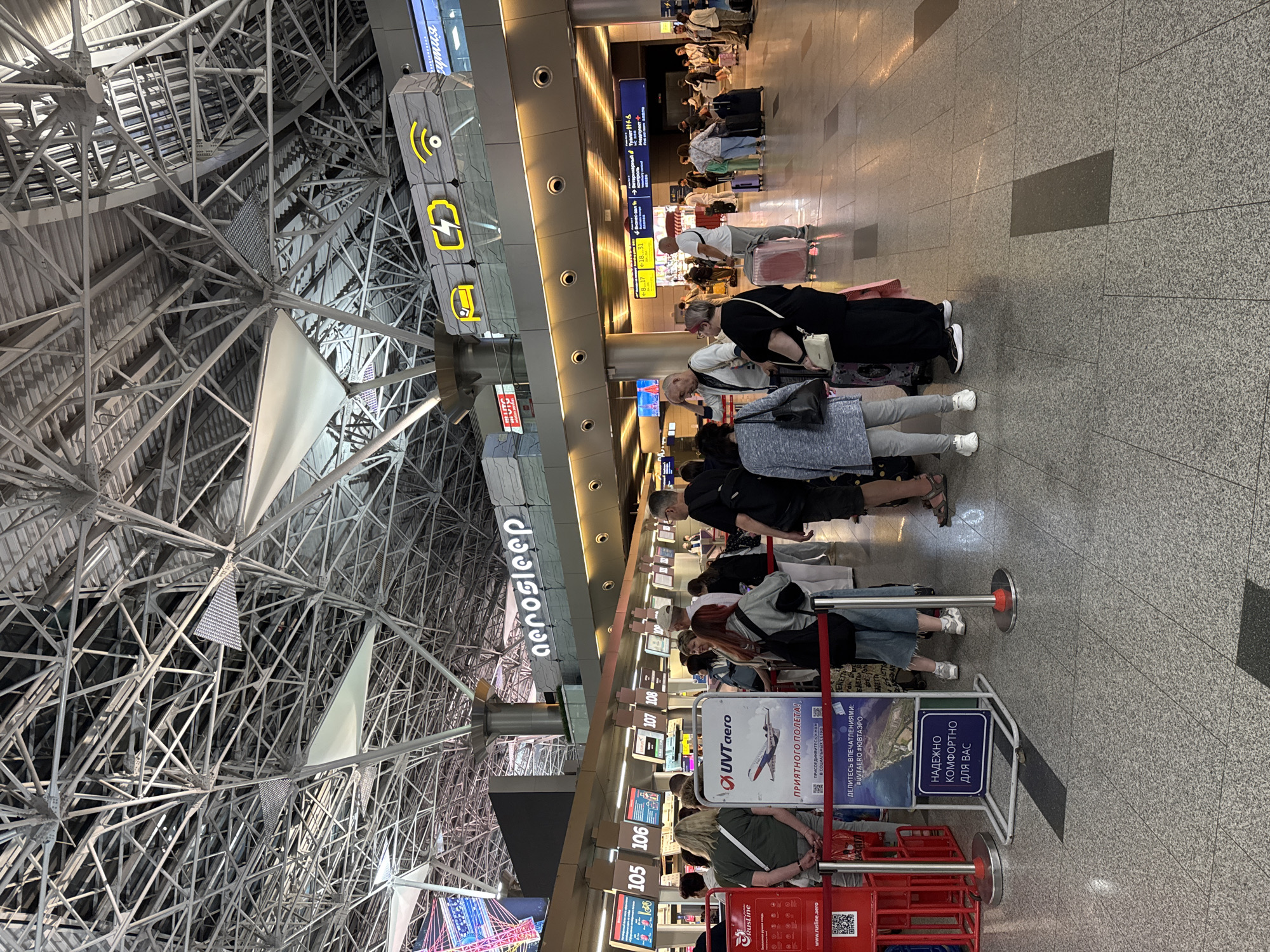 The image shows a bustling airport check-in area. A group of people are standing in line at various counters labeled with numbers such as "105," "106," "107," and "108." Above the counters, a sign reads "aerosleep." There are also signs indicating flight information, including one with yellow icons for a car, a lightning bolt, and a WiFi symbol. A prominent sign in the foreground, in Russian, says "НАДЕЖНО КОМФОРТНО ДЛЯ ВАС" which translates to "RELIABLE COMFORTABLE FOR YOU." Another sign with the "UVT aero" logo and an airplane graphic is also visible, along with text in Russian, including "ПРИЯТНОГО ПОЛЕТА!" or "HAVE A PLEASANT FLIGHT!". The airport ceiling features a complex, industrial metal truss structure. People are dressed in casual clothing and many have luggage. The floor is a grey tiled surface. The scene appears to be a busy departure hall at Vnukovo International Airport in Moscow, Russia, as suggested by the presence of Russian signage and the airport environment.