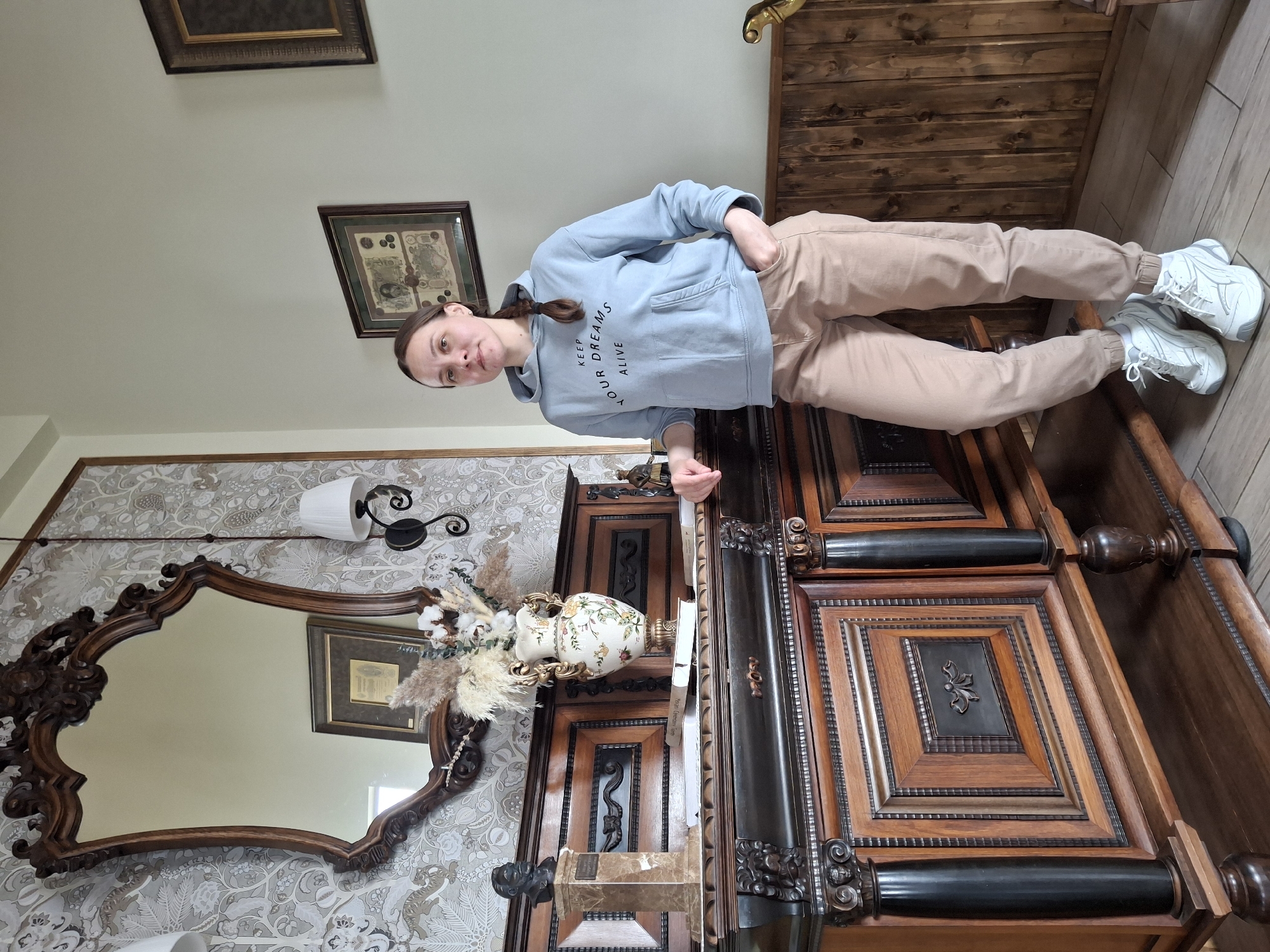 An indoor portrait features a young woman standing beside ornate dark wood furniture in what appears to be a room in Sarapul, Russia. The woman, with her hair in a braid, wears a light blue hooded sweatshirt with the text "KEEP YOUR DREAMS ALIVE" and light brown joggers, paired with white athletic shoes. She is looking directly at the camera with a neutral expression, one hand in her pocket and the other resting on the top of the large wooden cabinet or armoire.
The room's decor includes light-colored wallpaper with an intricate botanical pattern, a large, ornate wooden mirror on the left, and a decorative wall sconce above the mirror. On top of the wooden furniture, there is an ornate vase filled with dried flowers and pampas grass. Two framed pictures are visible on the upper walls, one of which has a distinct, possibly antique, design. The floor is made of wooden planks. The furniture itself is richly detailed with carvings, varying wood tones, and decorative black accents, suggesting a vintage or classic aesthetic. The overall lighting suggests it is daytime.
