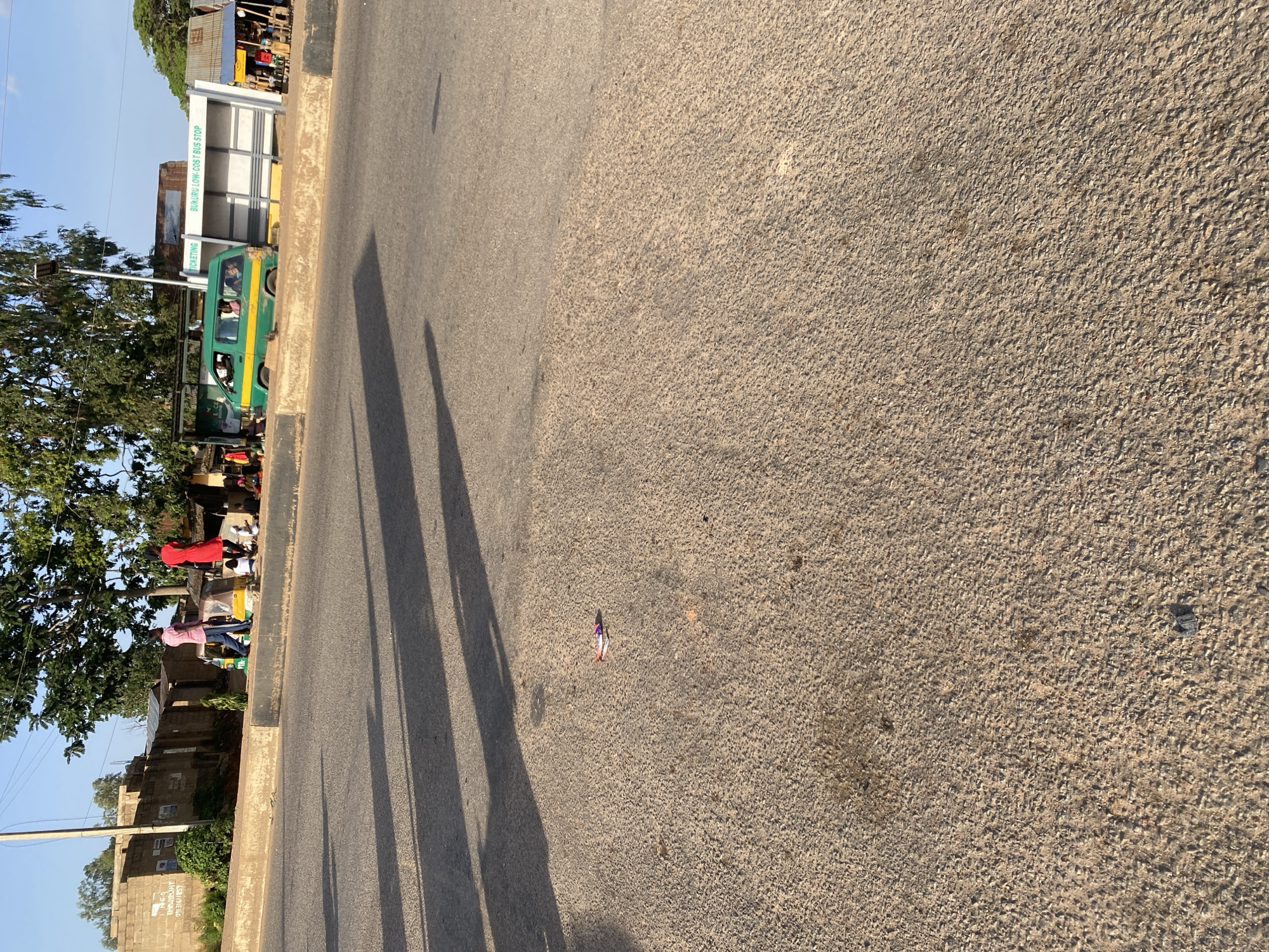 This image depicts a street scene at a bus stop in Jos, Nigeria. In the foreground, the rough asphalt of the road is visible, with a few scattered pieces of debris. In the middle ground, a beige concrete barrier separates the road from the sidewalk area. On the sidewalk, a green and yellow mini-bus is parked, labeled "TICKETING" and "BUKURU LOW-COST BUS STOP." Several people are gathered around the bus and the bus stop shelter, some standing and some sitting. Two individuals, one in a pink shirt and blue pants and another in a red dress, are walking away from the viewer. Behind the bus stop, a brick wall and some greenery are visible. The sky above is clear and blue.