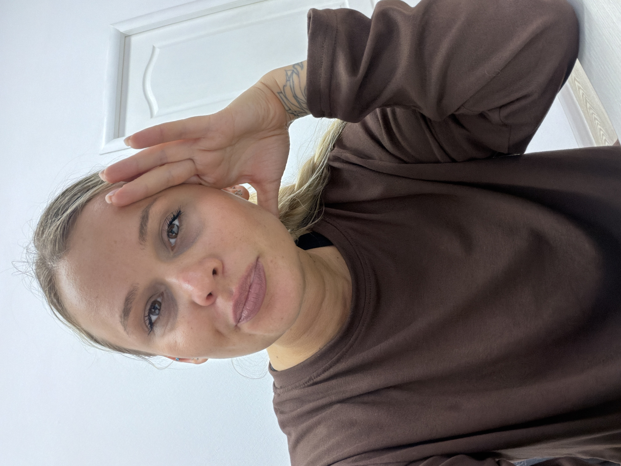 A woman with blonde hair pulled back and wearing a brown long-sleeved shirt is looking down at the camera, her right hand on her forehead. The background is a white wall with a white door in the upper left corner. The location is not specified in the image, and therefore cannot be attributed to Sarapul, Russia.
